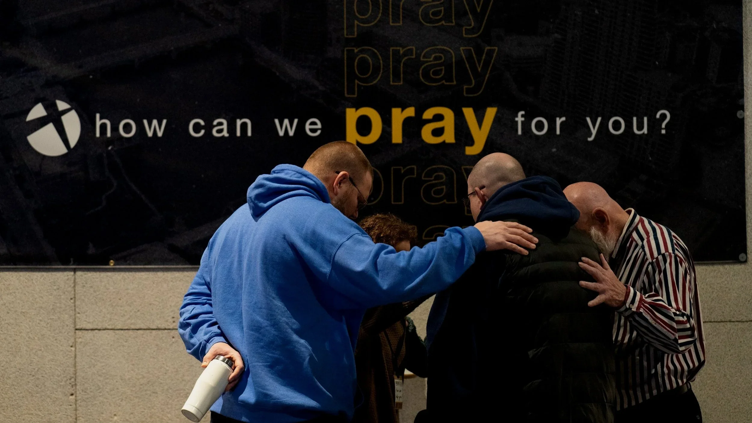 Group of people praying together with their heads bowed and hands placed on each other's shoulders in front of a black sign that reads 'how can we pray for you?' with a church logo.