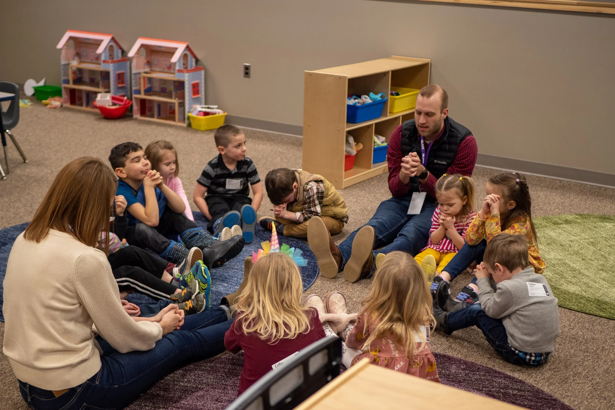 Children sitting on the floor in a classroom, engaged in an activity led by an adult.