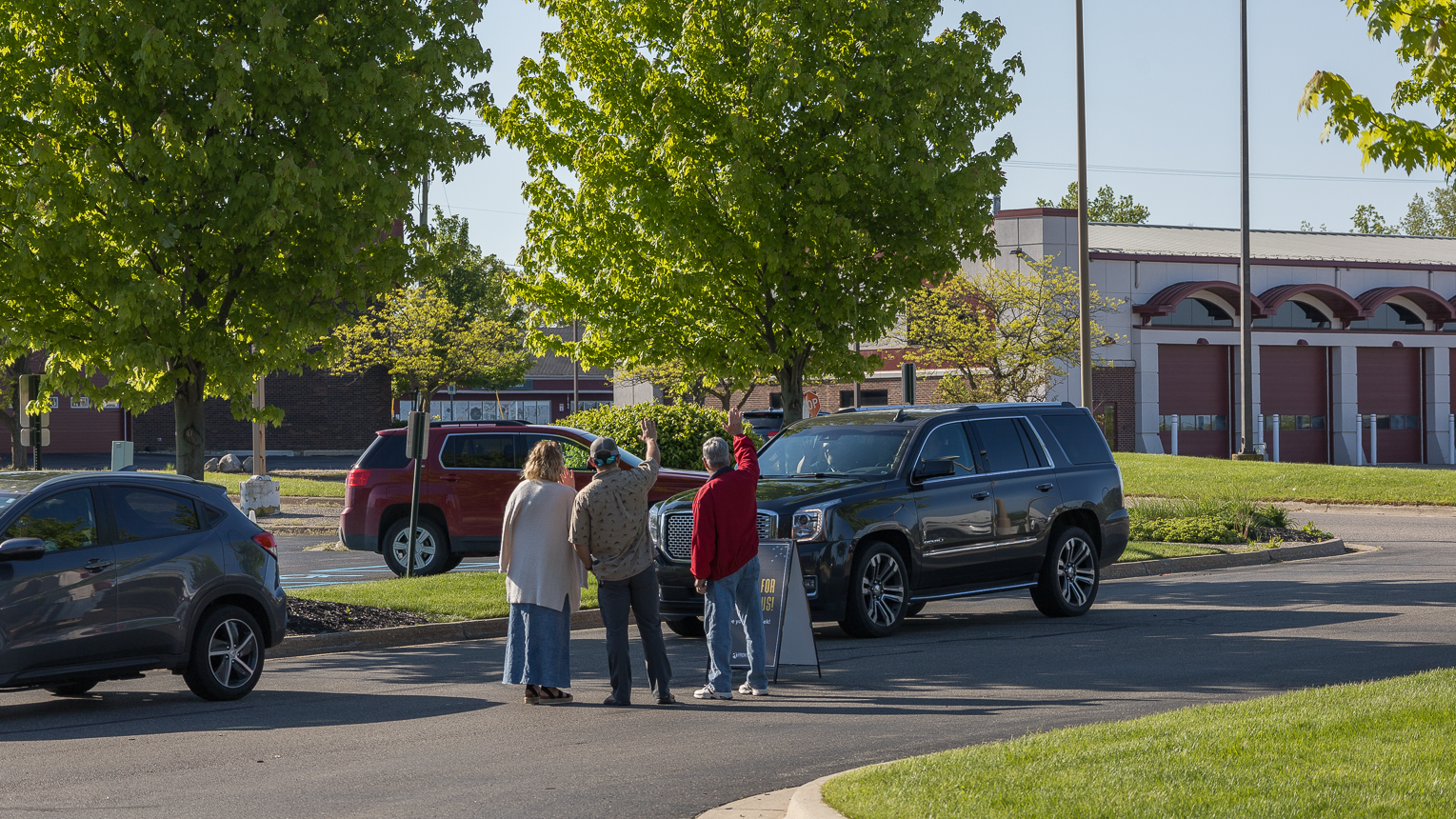 Three people standing in a parking lot near a black SUV, having a conversation and waving, with trees, other parked cars, and a building in the background.