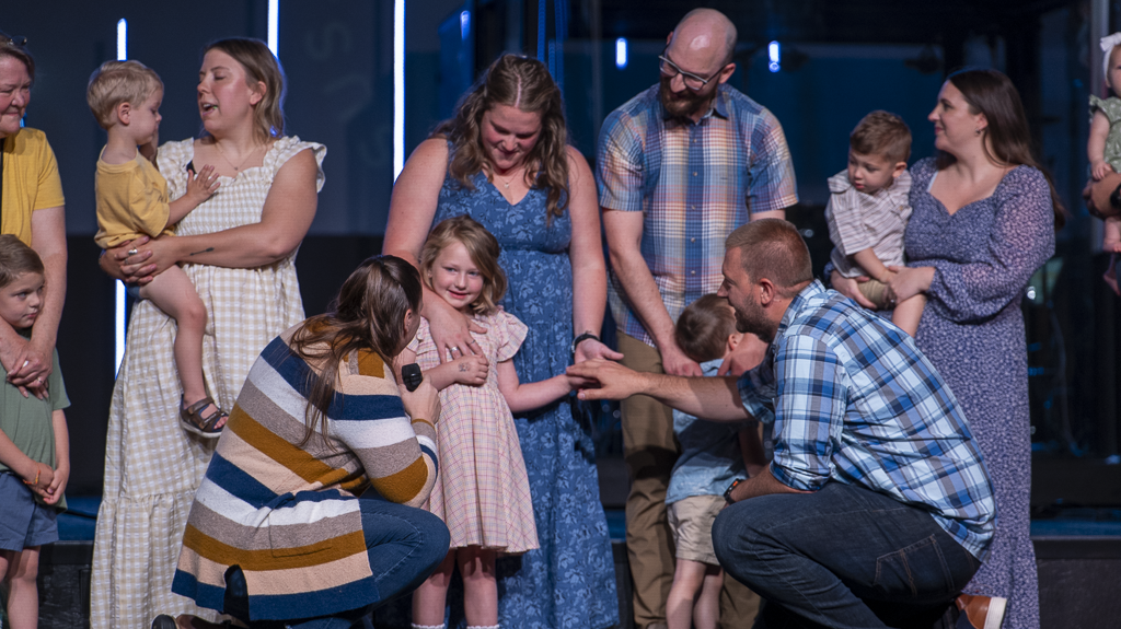 A group of adults and children gathered on stage, with a woman holding a microphone kneeling and talking to a young girl in a pink dress. Others are smiling and engaging as part of a church or community event.