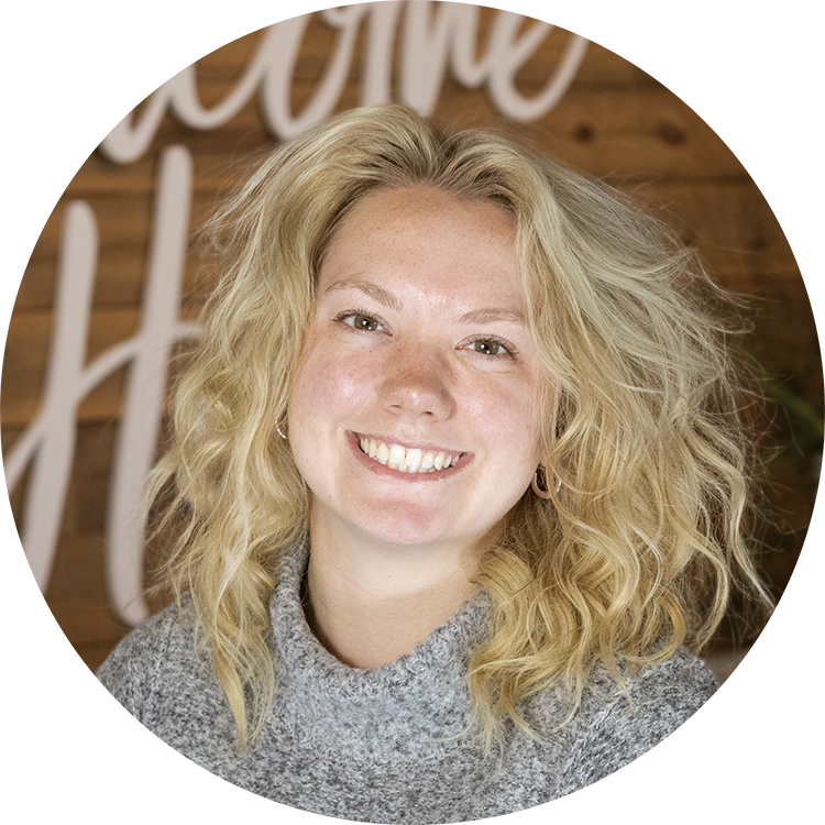 Smiling blonde woman with curly hair wearing a gray sweater, standing in front of a wooden wall with white cursive writing.