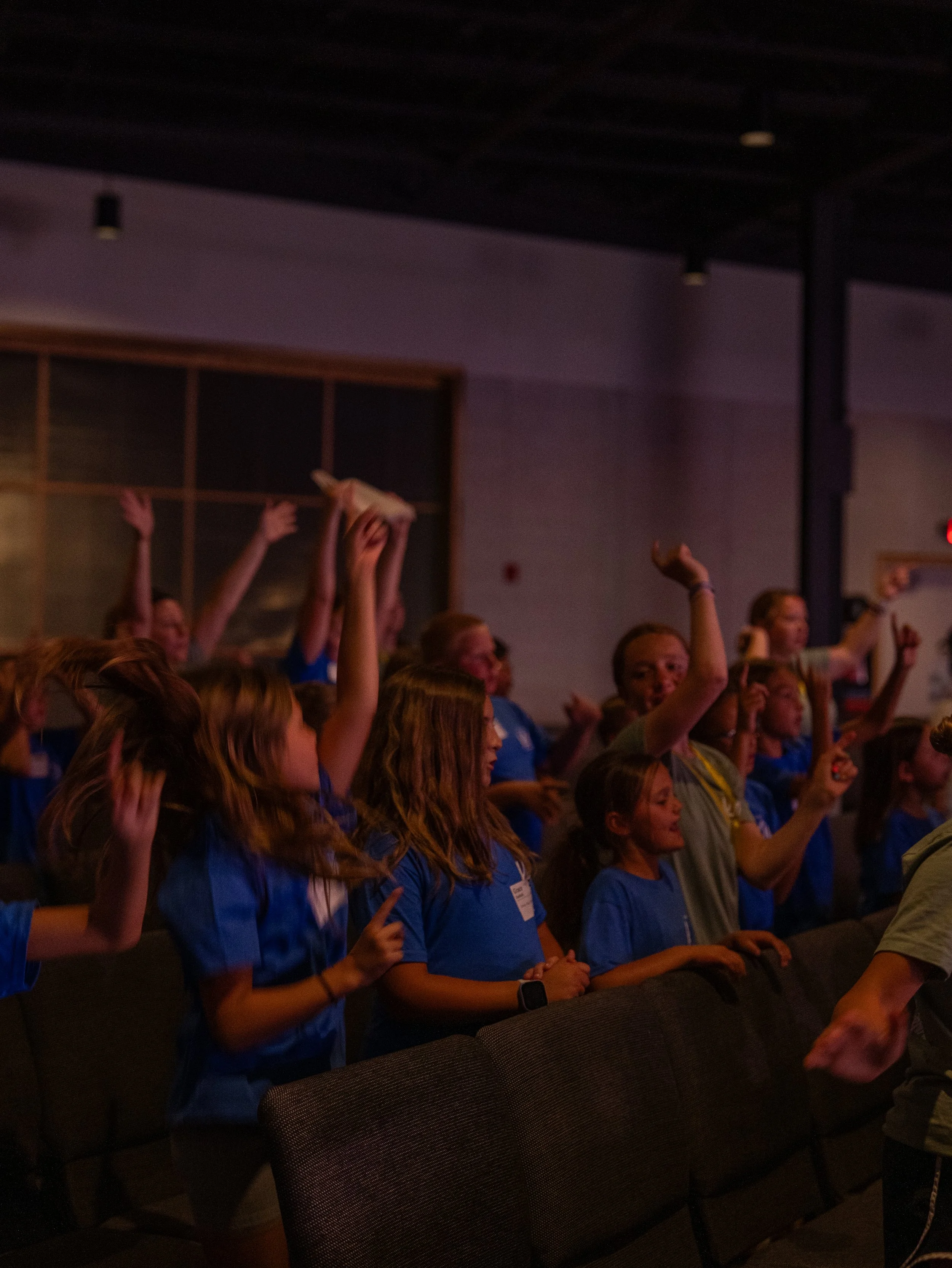 A large group of young people, most wearing blue shirts, raising their hands in a dark indoor setting, possibly participating in a group activity or celebration.