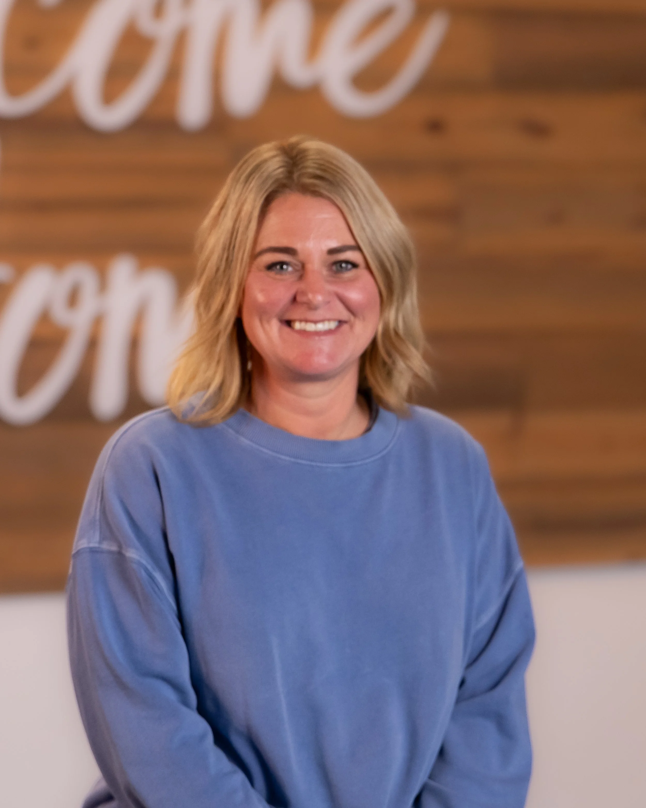 A smiling woman with blonde hair sitting in front of a wooden wall with the words 'welcome home' in cursive. Potted plants are visible in the background.