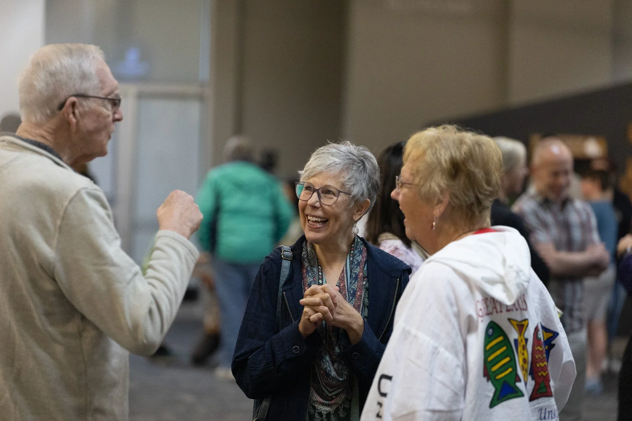 A group of older adults, including three women and one man, engaged in conversation at an indoor event. Two women are smiling and holding hands, while the man and another woman participate in the discussion. The background shows other attendees and a large room.