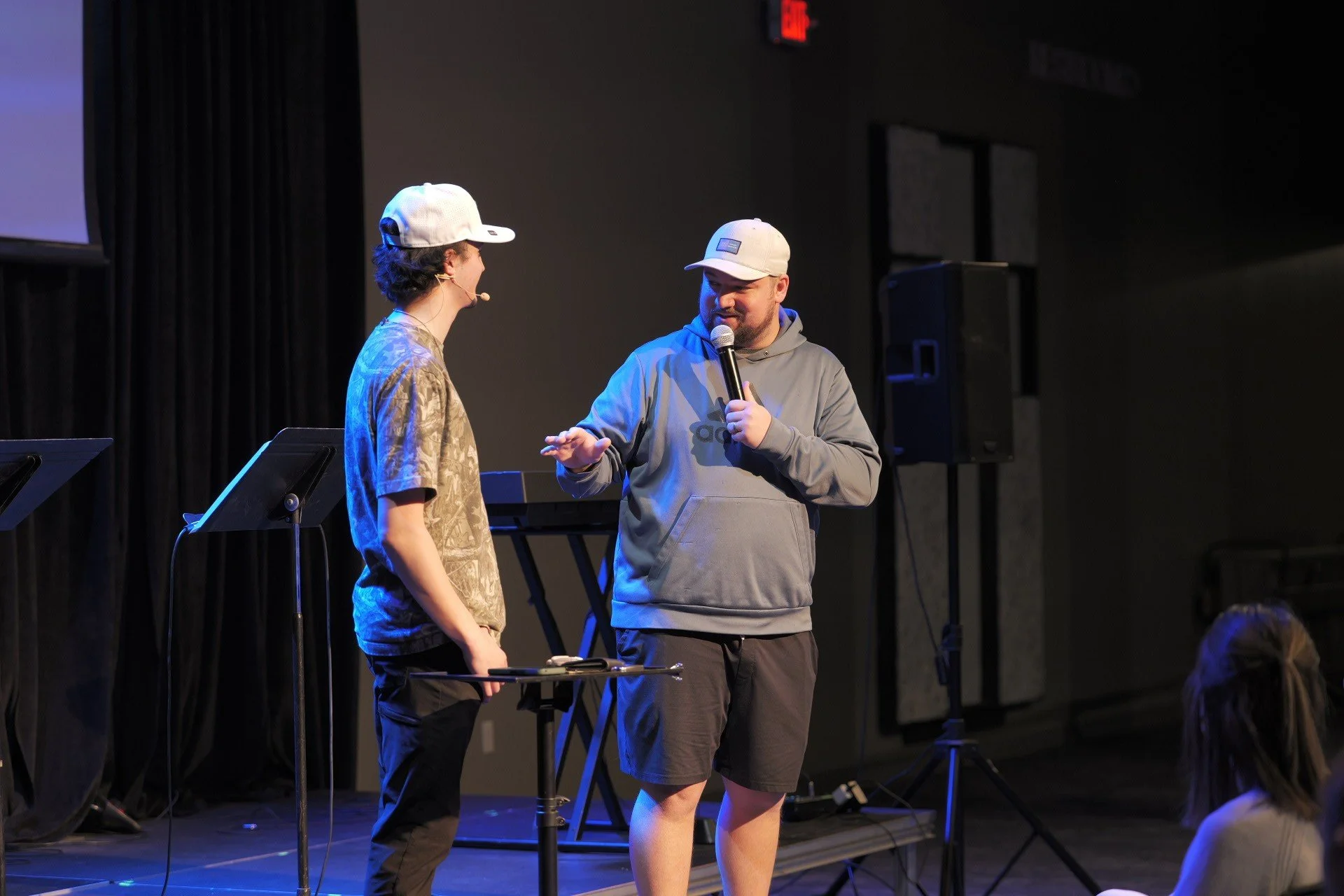 Two men having a conversation on stage, both wearing gray hats and casual clothing. One is holding a microphone and smiling, while the other is listening.
