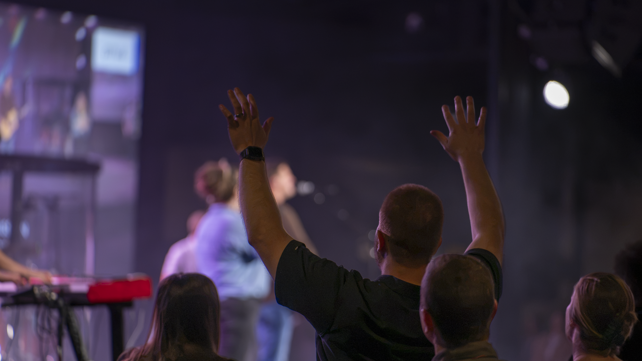 Audience with raised hands at a live event or concert, with stage lights and performers in the background.