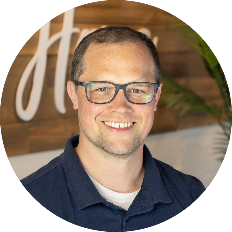 A smiling man with short brown hair, glasses, wearing a navy blue shirt, standing indoors with a wooden wall and a leafy plant in the background.