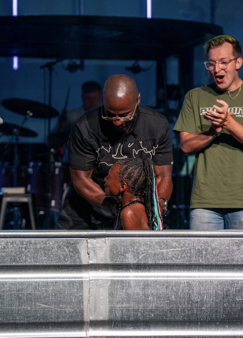A young girl with braided hair is in a baptismal pool, being baptized by a man wearing glasses and a black shirt. Two other men are standing nearby, one holding a phone. The background shows a stage with drums and music equipment.