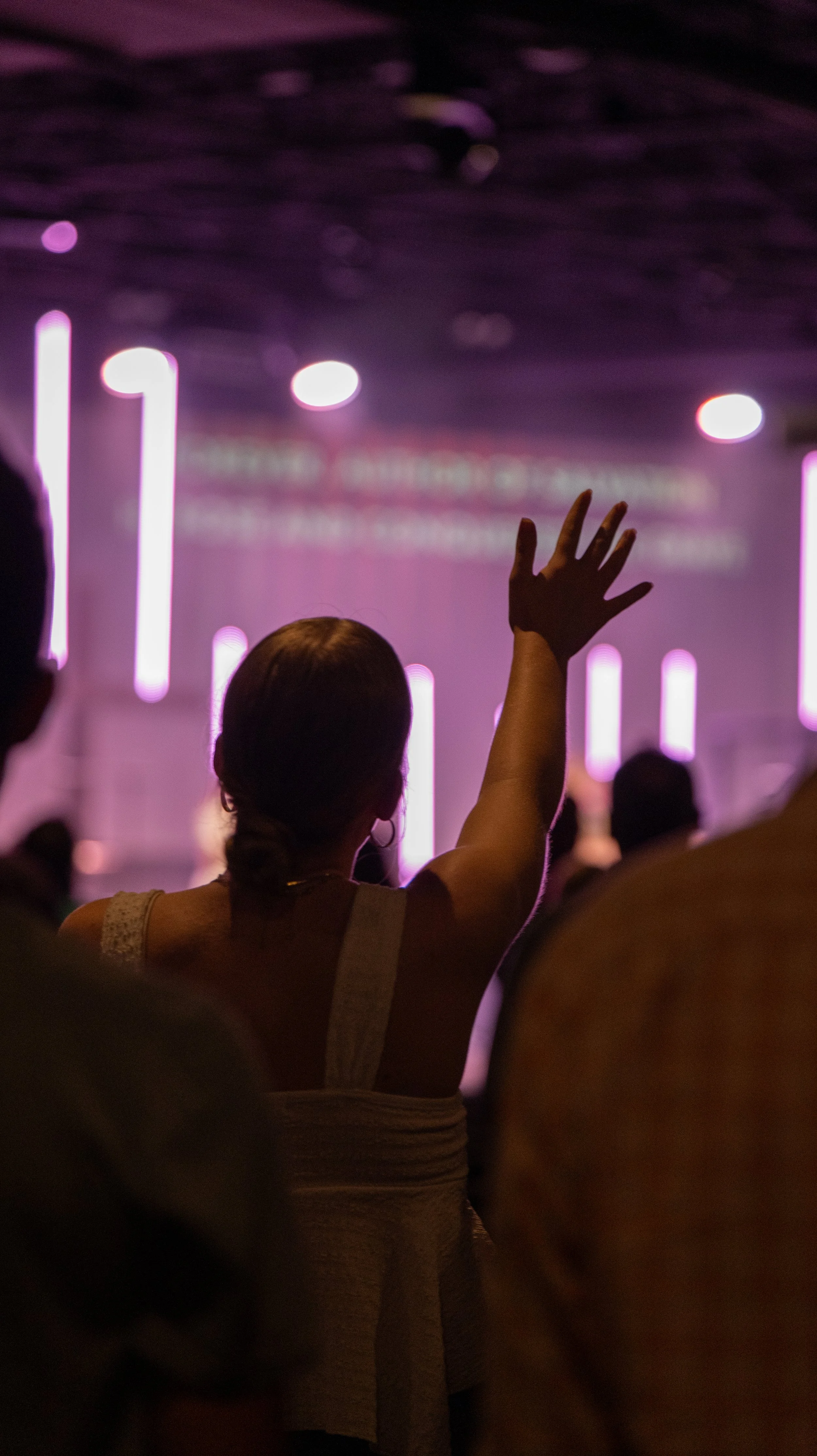 A woman raising her hand in a crowd at an indoor event with purple lighting and blurred background.