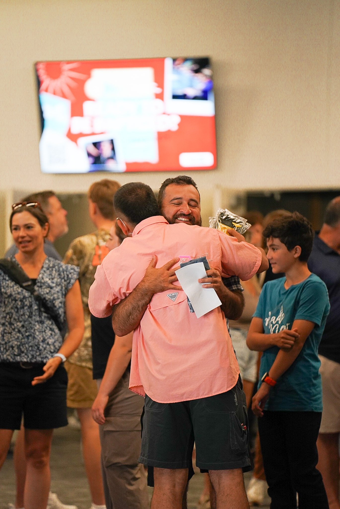People gathered in an indoor setting with a large screen on the wall, engaging in a hug and conversation, with some smiling and others observing.