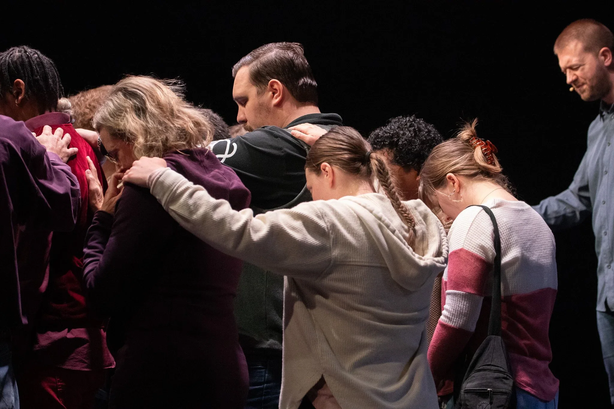 A group of people praying together with their heads bowed and hands placed on each other's shoulders or heads in a circle.
