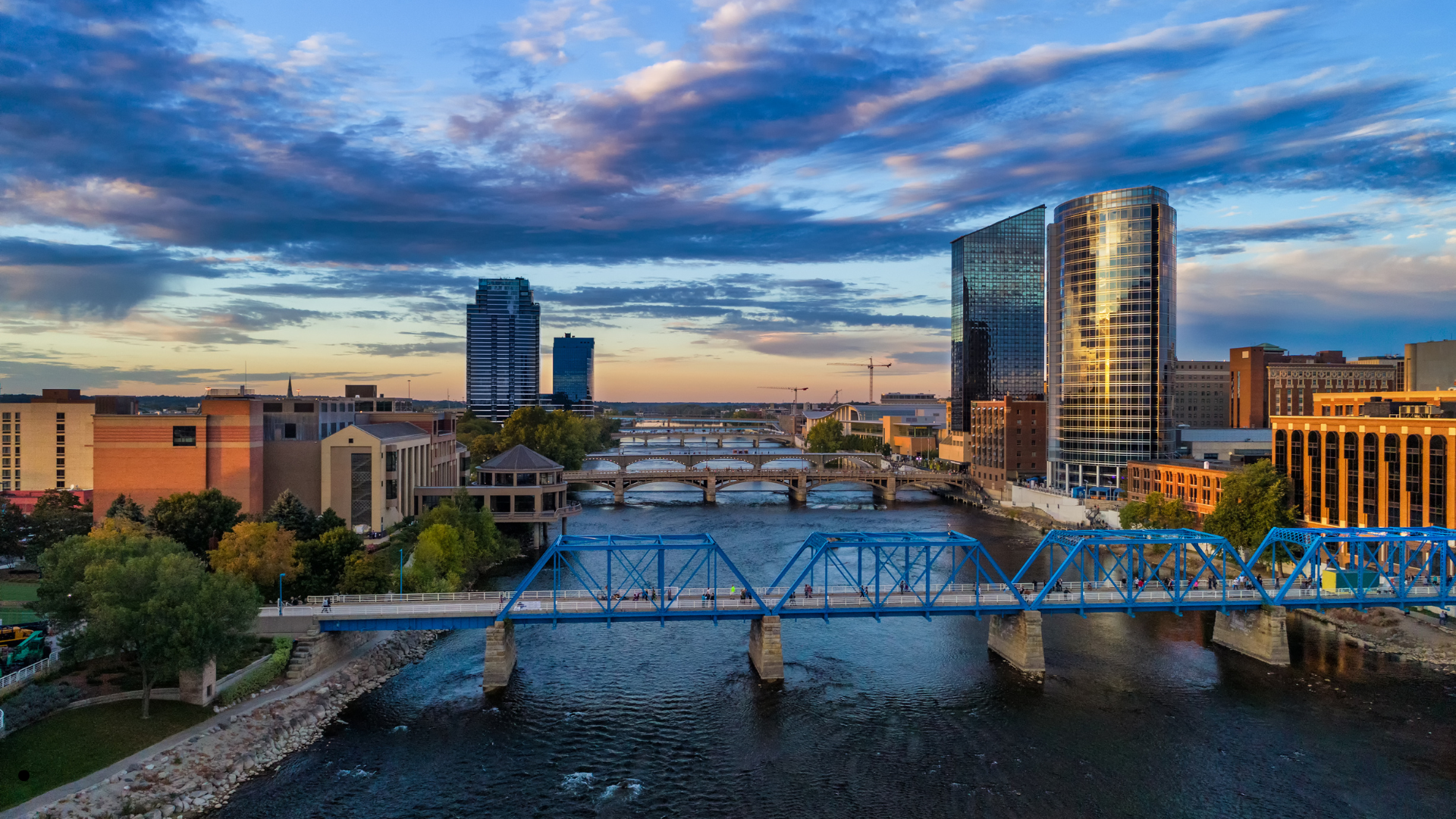 City skyline at sunset featuring modern glass buildings, a river with multiple bridges, and a partly cloudy sky.