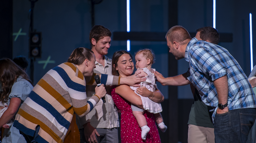 Group of people on stage, including a woman holding a young child, impacting or blessing her, with others assisting or participating.