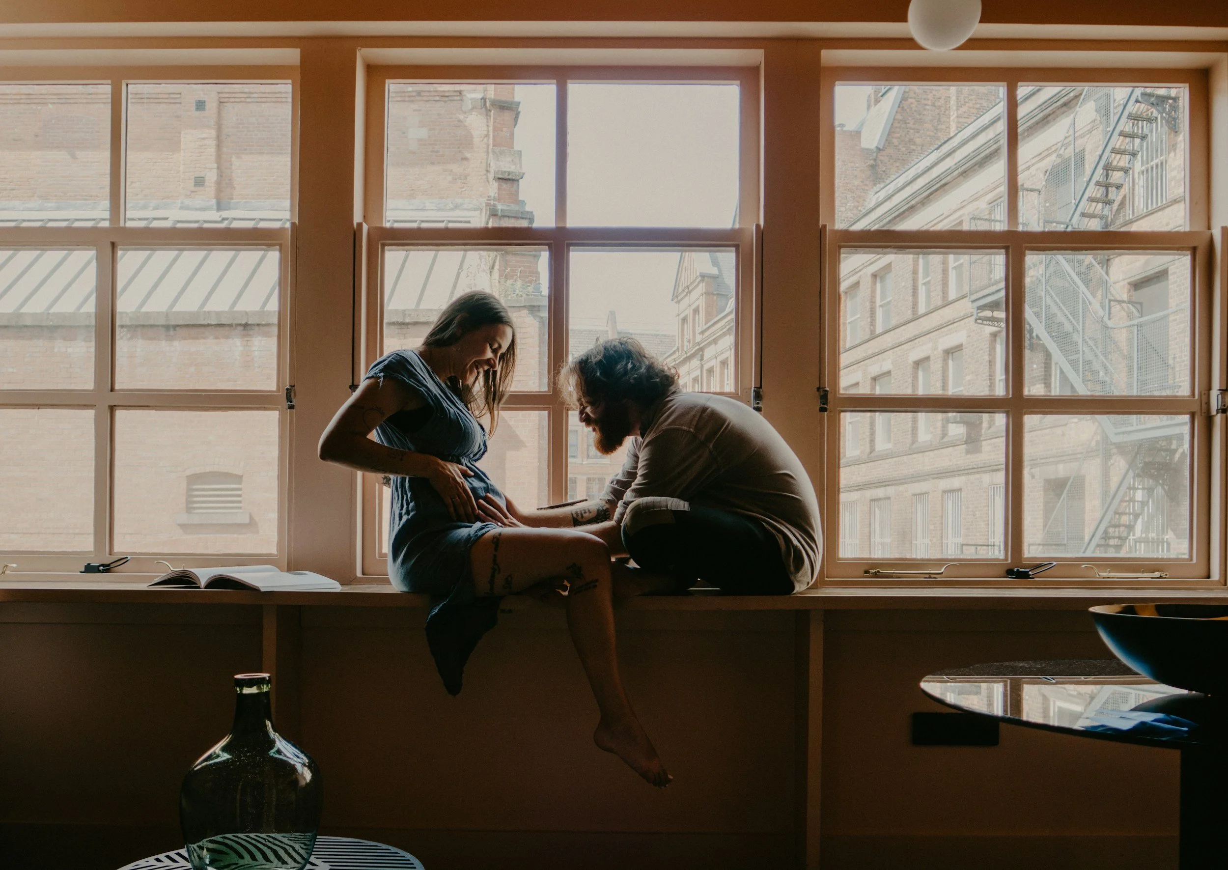 A couple sits on a wide window sill inside a room, with large windows showing an urban brick building exterior outside. The woman, visibly pregnant, smiles and looks down at her belly while the man leans in, gently touching her belly. There is a book open on the window sill and a large decorative vase on a table below.