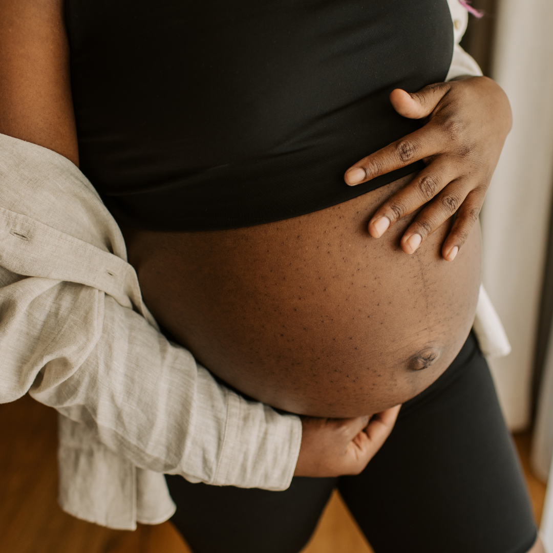 Close-up of a pregnant woman with dark skin, wearing a black top. Someone's hand rests on her pregnant belly, which has visible stretch marks, as part of a beige shirt sleeve is visible.