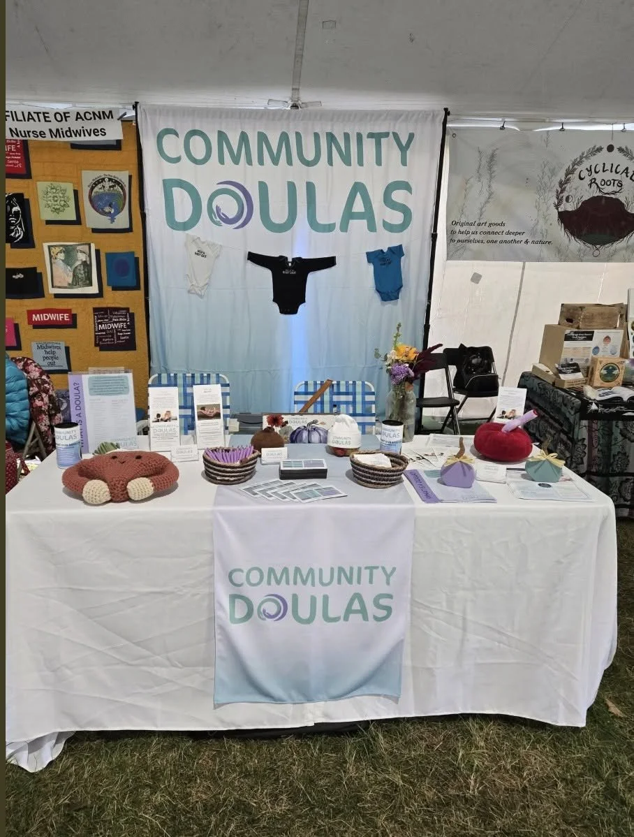 A booth with a banner that reads 'Community Doulas' featuring prenatal and baby items, informational pamphlets, and decorative handmade crafts like knitted baby hats and stuffed toys.