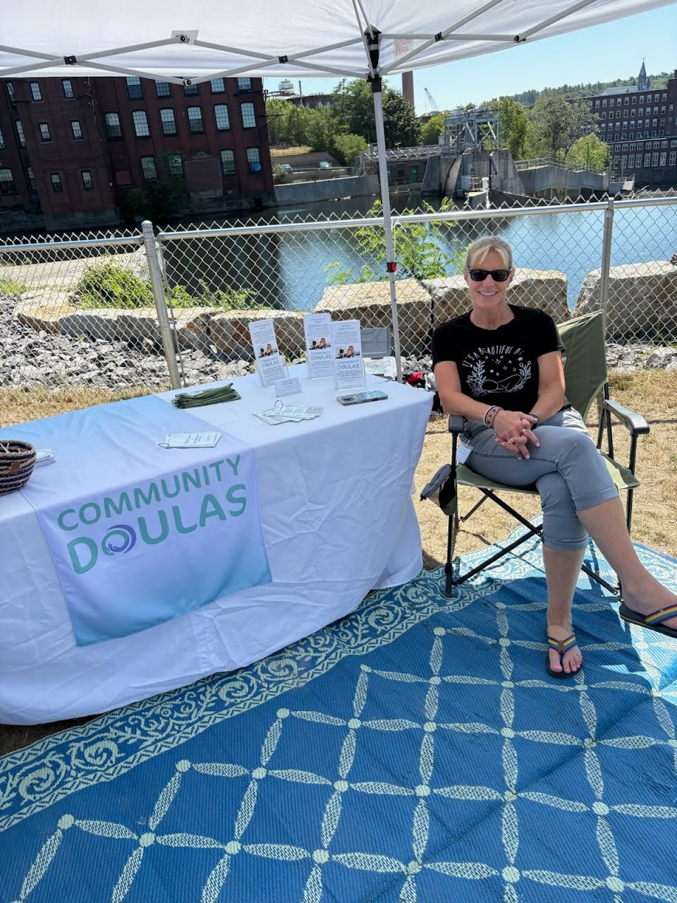 Woman sitting by a table with 'Community Doulas' banner outdoors near a river, with an umbrella overhead and buildings in the background.
