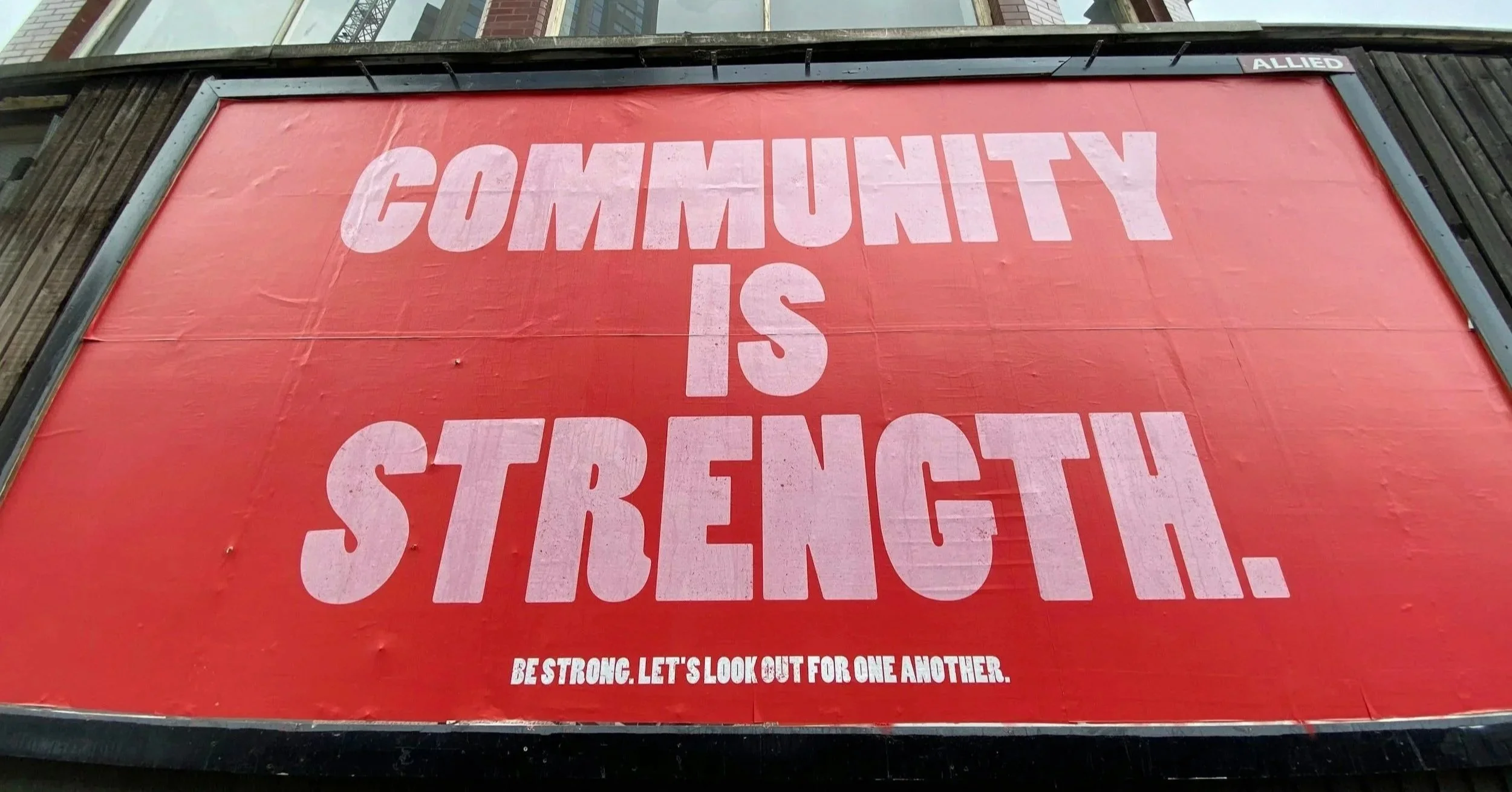 Red sign with white text that reads, 'Community is Strong.' The sign is mounted on a building, with a small caption at the bottom that says, 'Be strong. Let's look out for one another.'