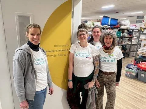 Four women standing inside a facility, wearing matching Community Doulas volunteer t-shirts, smiling for the photo.