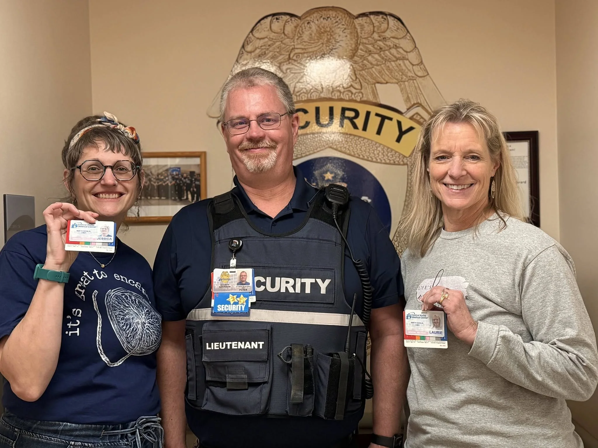 Three smiling people standing close together in front of a sheriff's badge emblem on the wall. The woman on the left has glasses, and holds an ID badge. The man in the middle is a  security officer with short gray hair, glasses, wearing a uniform with a badge, and a vest labeled 'LIEUTENANT'. The woman on the right has long blonde hair, and is wearing a light gray sweater, holding a badge and smiling.