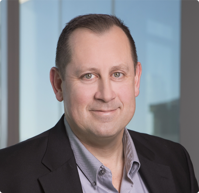 Business professional man wearing a black blazer and gray shirt, standing in front of a blurred office background.