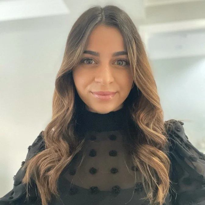 A woman with long, wavy brown hair and fair skin, smiling slightly, wearing a black, sheer top with a high neckline and decorative buttons, standing indoors with a light-colored background.