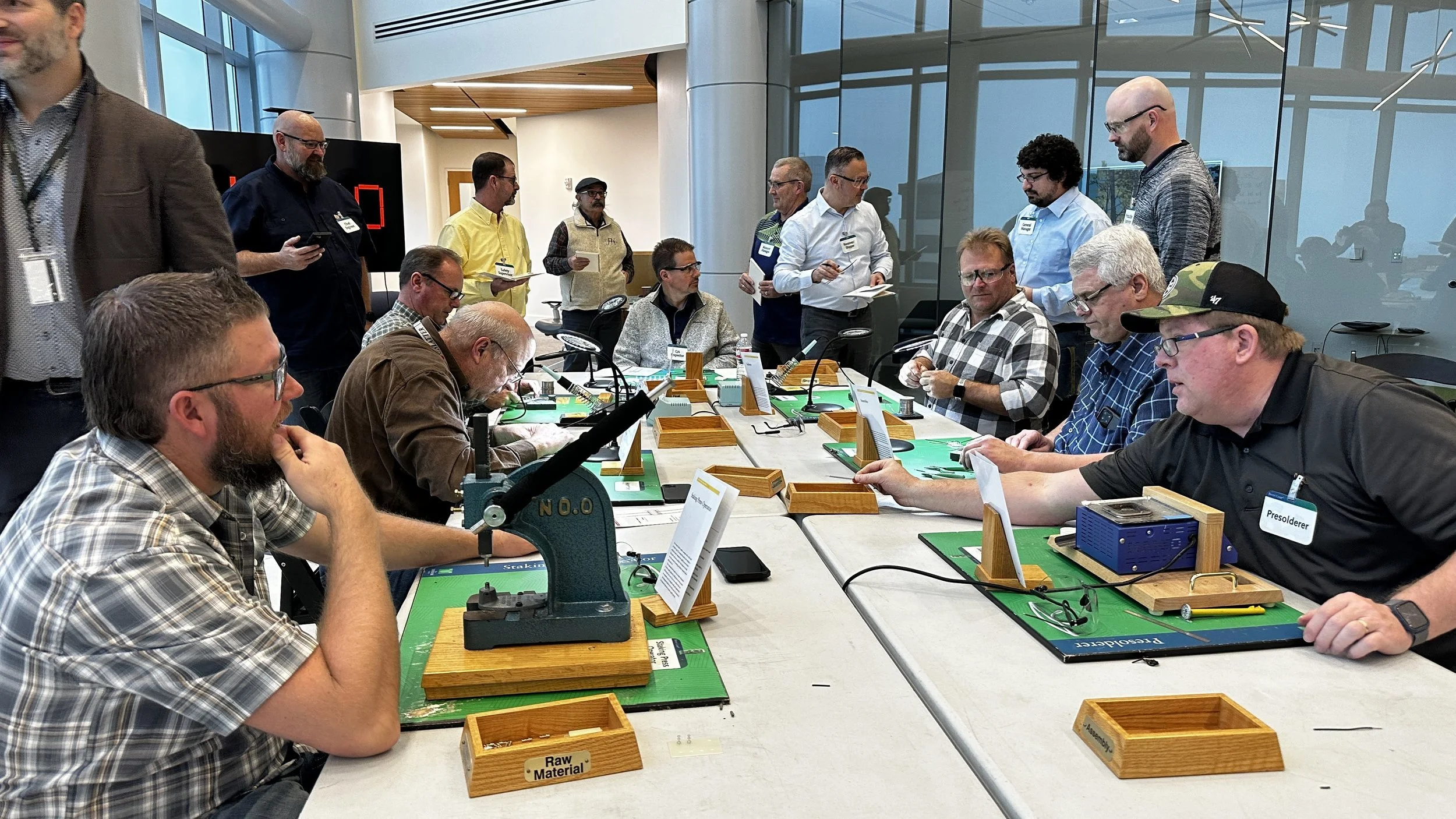People sitting at a long table engaged in a game or activity, with some standing around observing. The table has green mats, wooden boxes, and various objects related to the game or activity. The setting appears to be a modern indoor space, possibly 