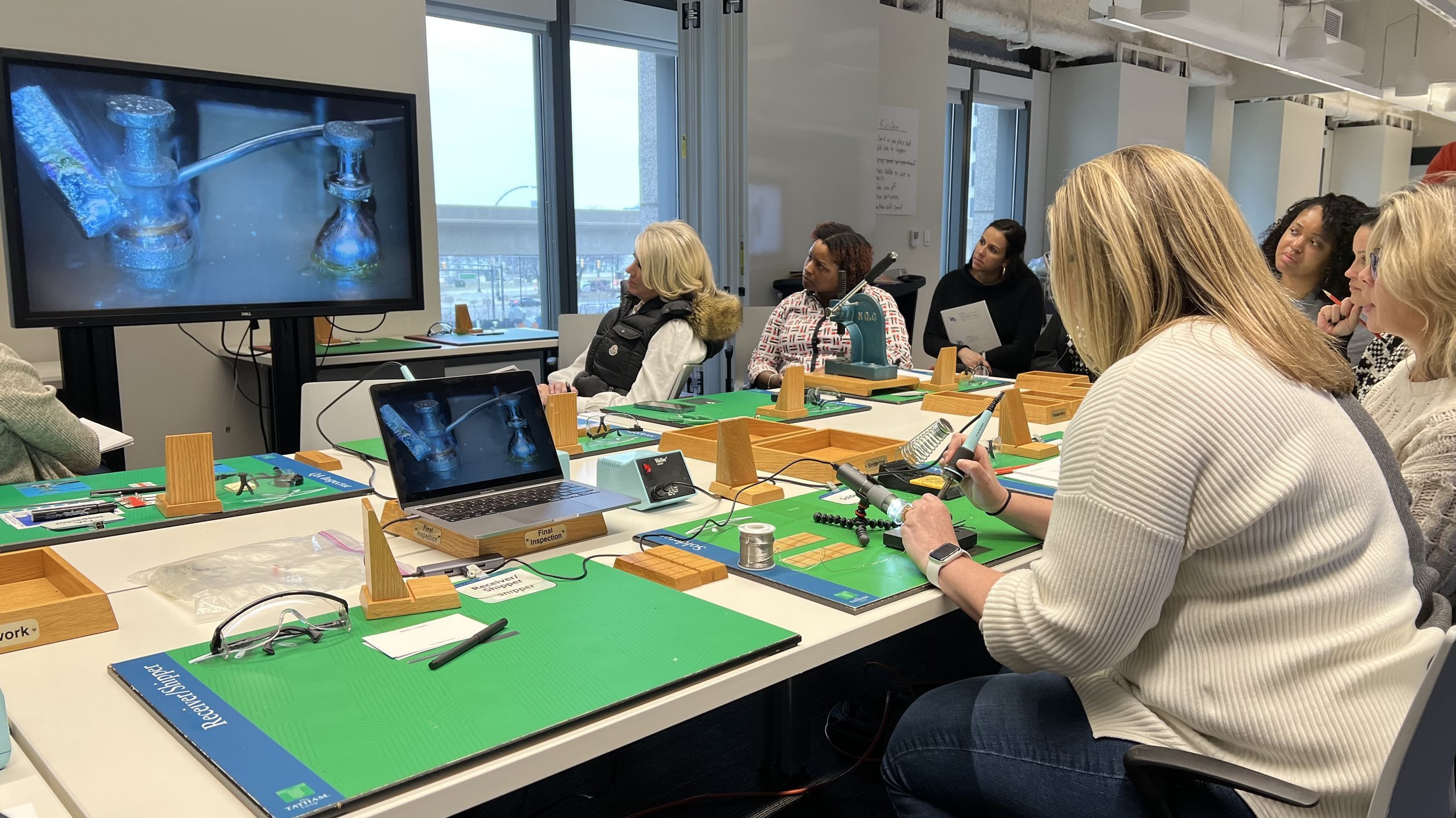 Group of women sitting at a table in a classroom or workshop setting, watching a demonstration of plumbing or faucet repair on a large screen. The table is equipped with work mats, tools, and educational materials.