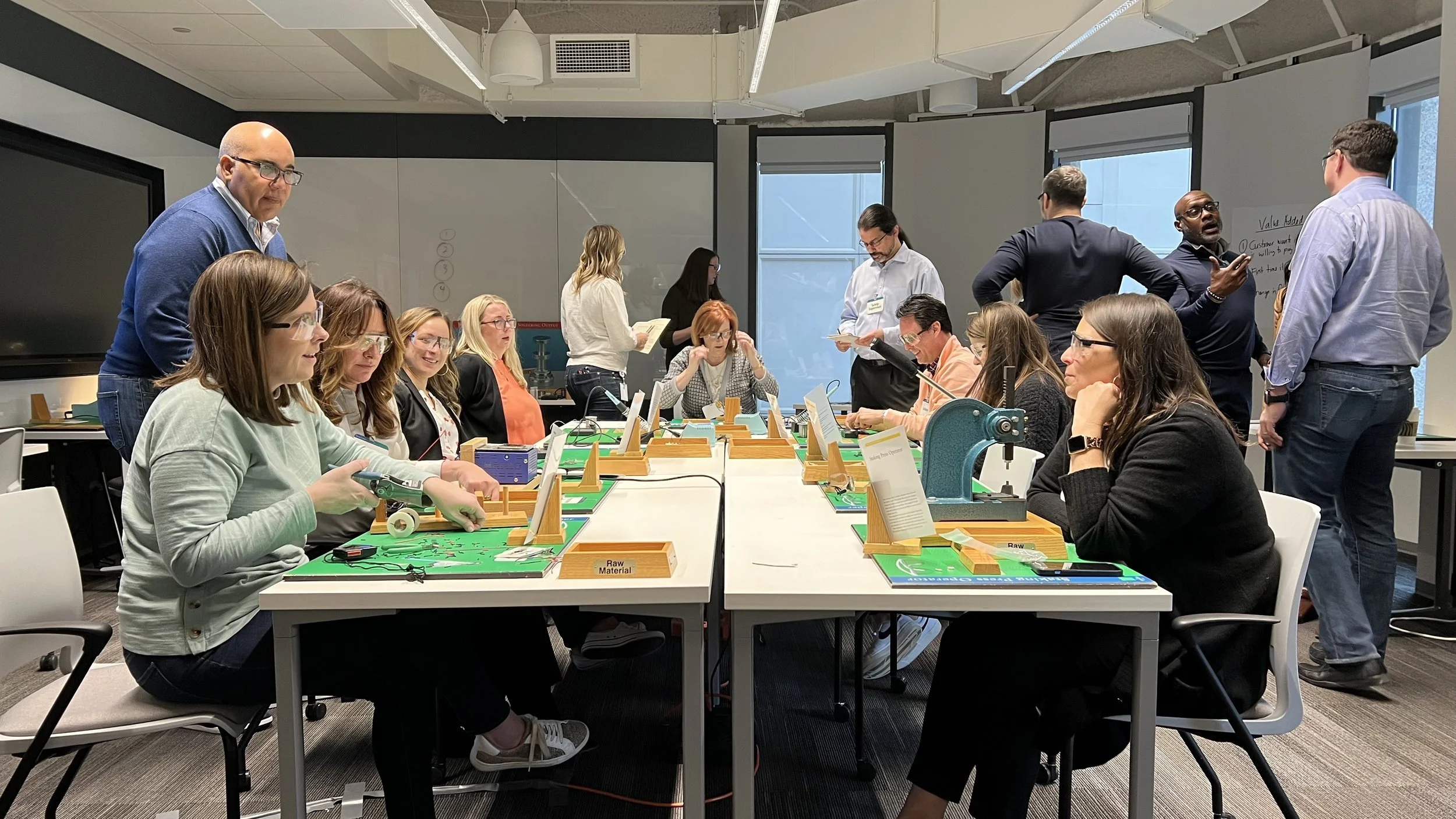 A group of adults in a meeting room engaged in a collaborative workshop. Some are seated around a table working with electronic devices, while others stand and discuss near a whiteboard. The room has large windows and modern furnishings.