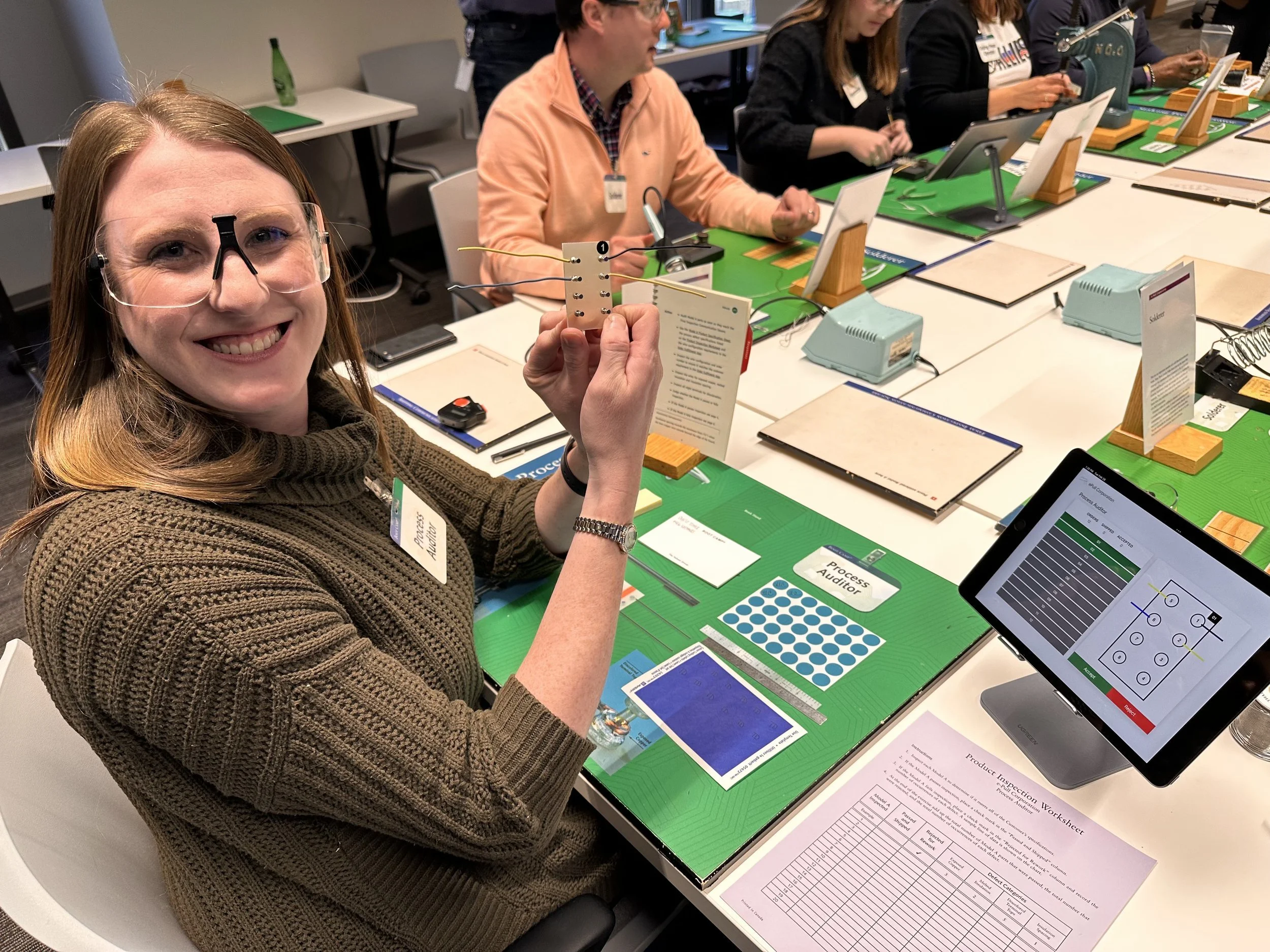 A woman smiling and holding a small circuit board in a conference room with multiple people working at a long table with tablets, documents, and electronic equipment.