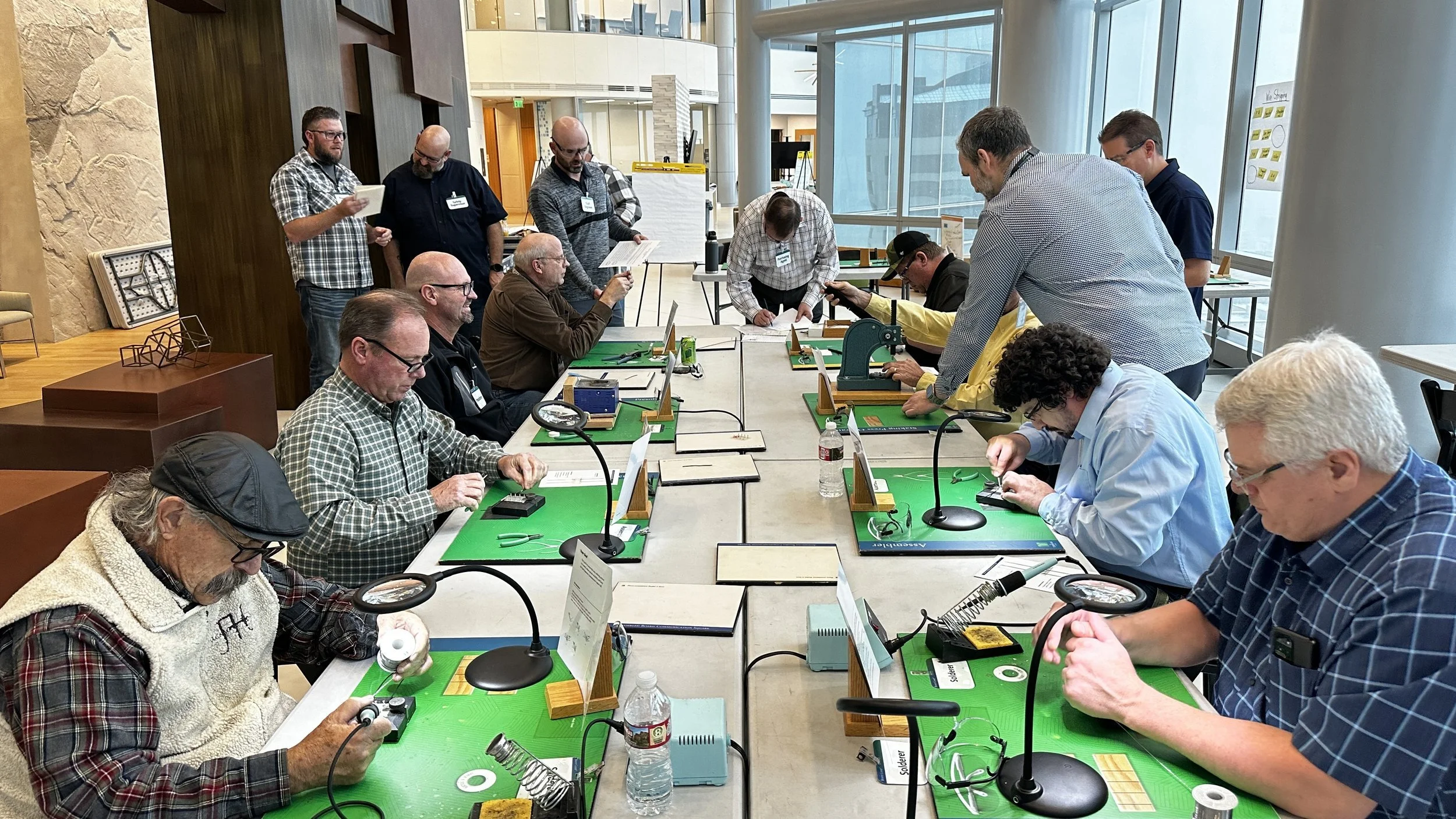 A group of people participating in a hands-on electronics workshop, working with soldering stations on green mats at a conference table, with some standing and others seated.
