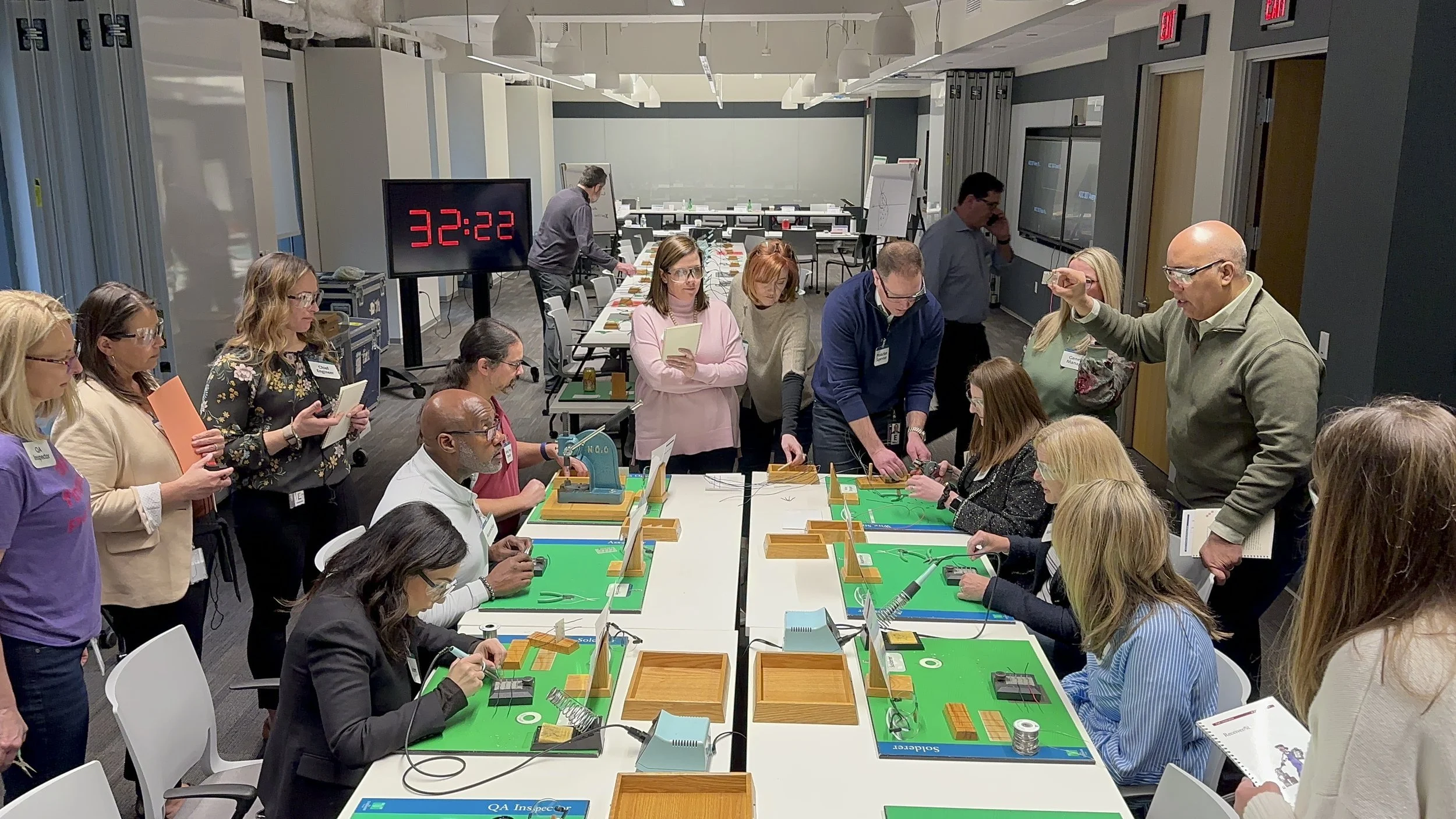 A group of people participating in a wire-connection workshop in a conference room. Some are seated at tables with green mats, working with tools, while others stand around observing. A man in a beige jacket is instructing, and a large digital clock displaying 32:22 is visible in the background.
