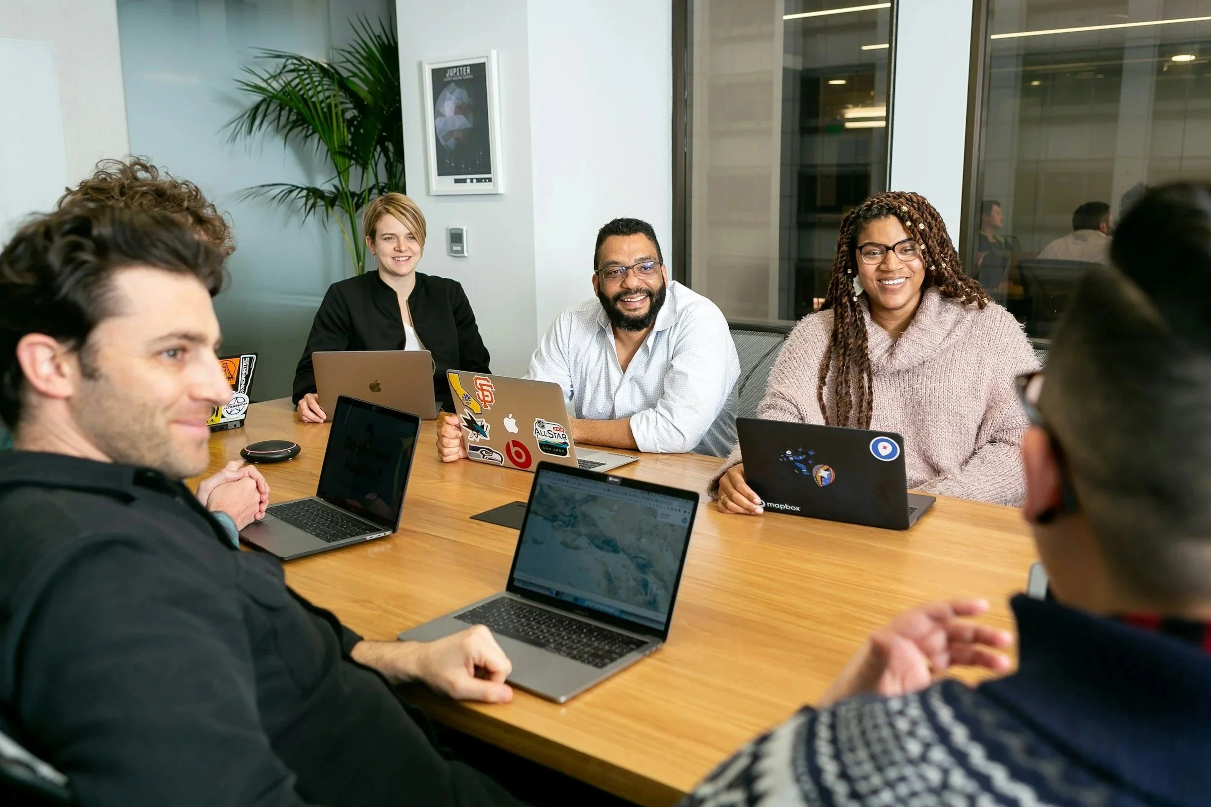 A diverse group of six people smiling and having a meeting around a wooden conference table with laptops in a modern office.
