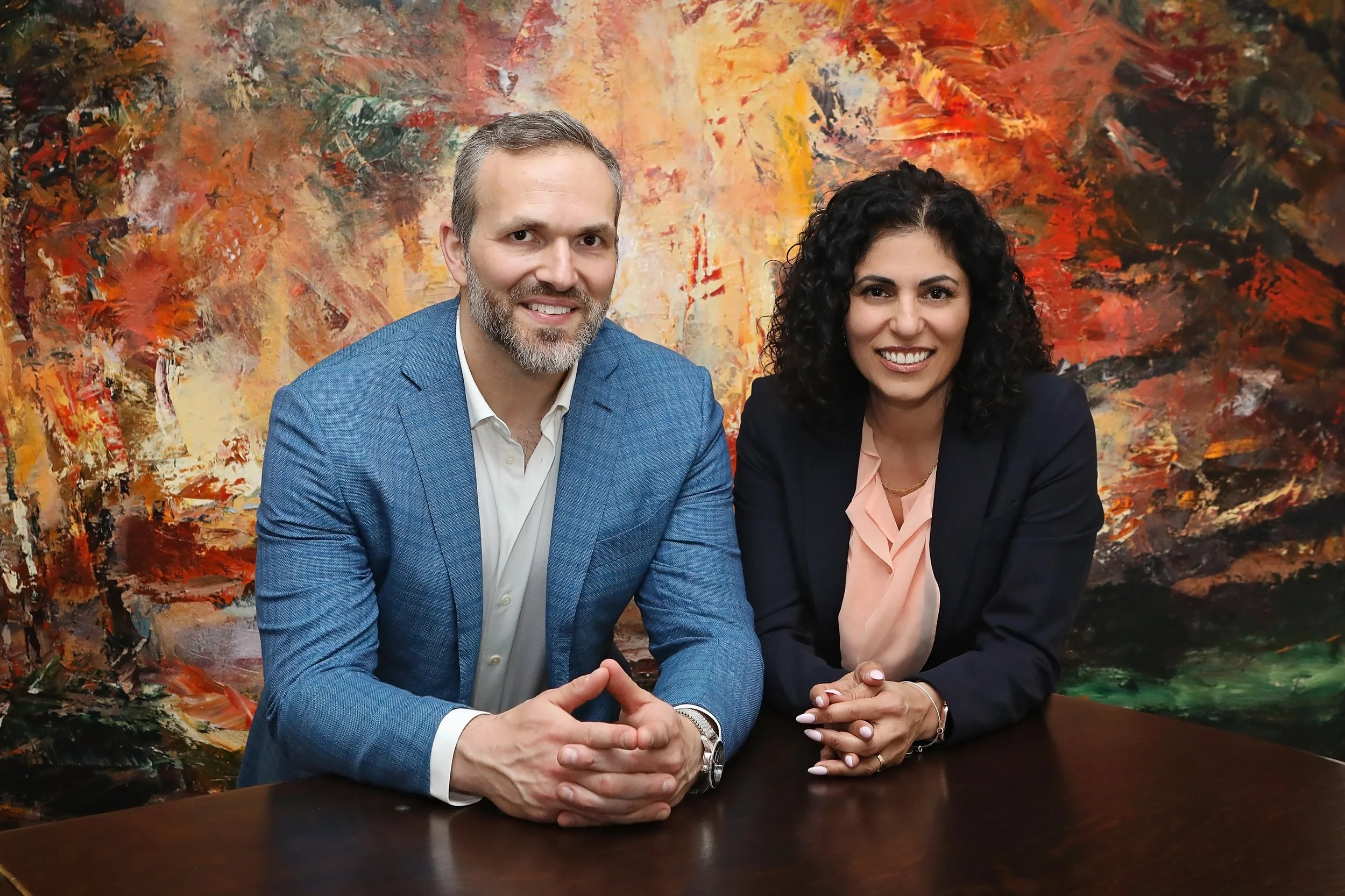 A man and woman sitting at a dark wood table in front of a colorful abstract painting. The man has a beard, gray hair, and wears a blue blazer with a white shirt. Both are smiling.