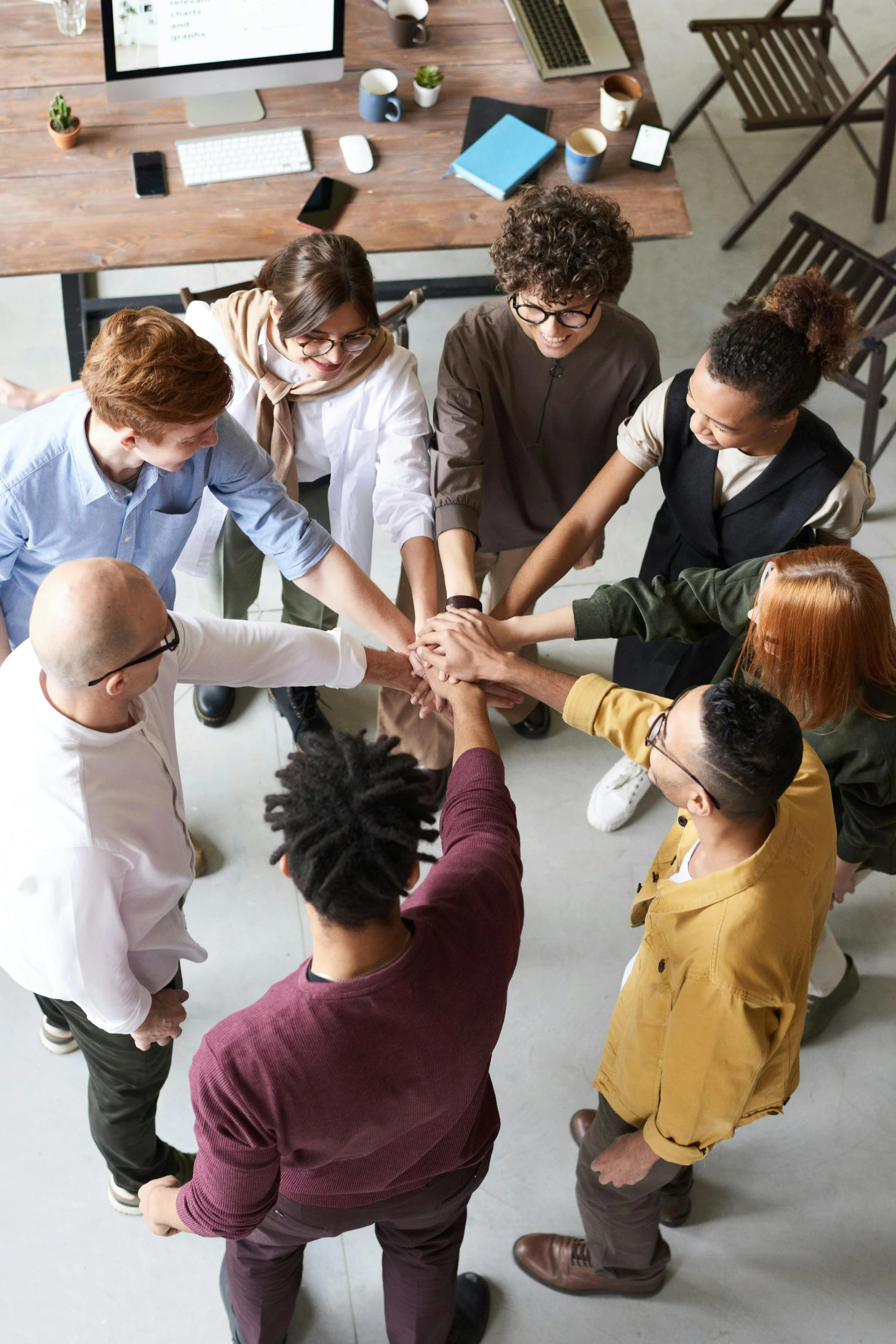 A diverse group of nine people standing in a circle with hands stacked in the center, smiling and showing teamwork and unity in a modern office space.
