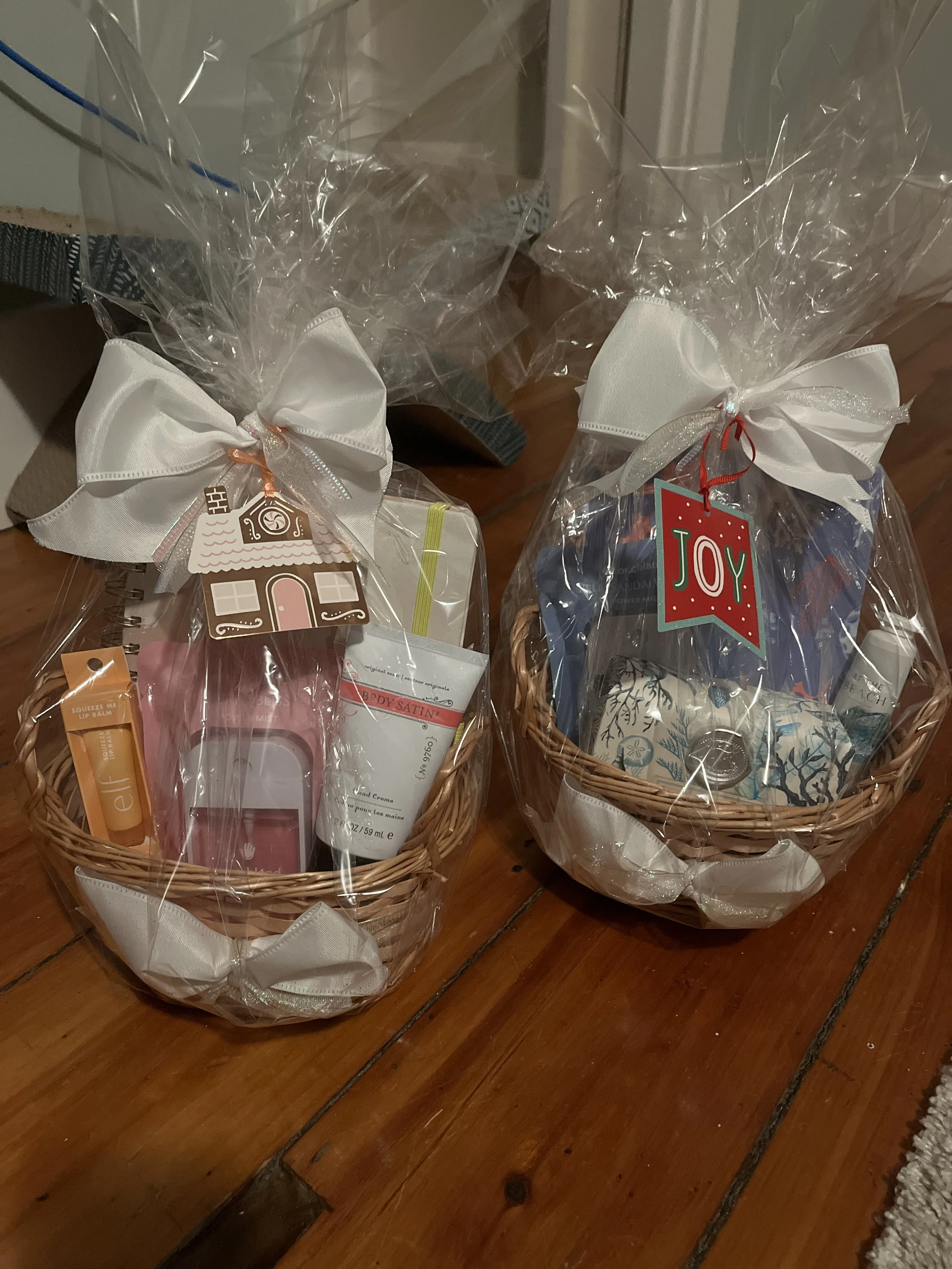 Two gift baskets wrapped in clear plastic with white ribbons and holiday tags, placed on a wooden surface.
