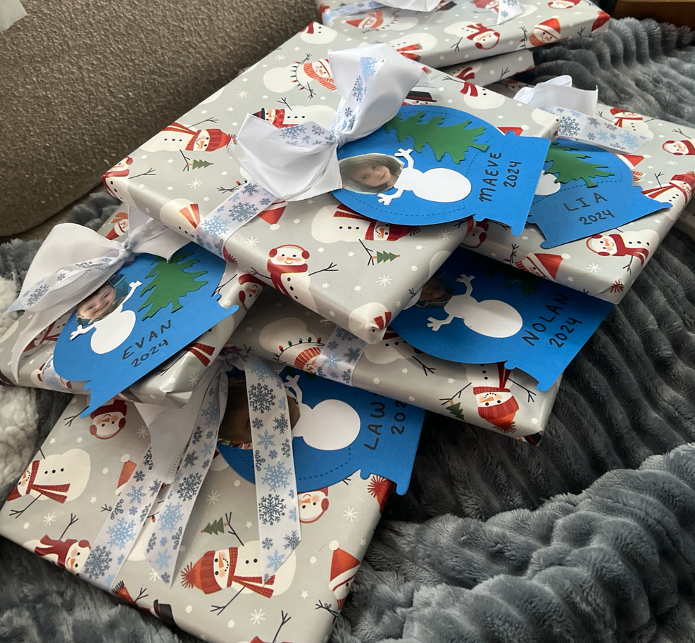 Several wrapped Christmas presents with snowman and holiday-themed wrapping paper. Each gift has a blue tag with a child's photo, name, and the year 2024, attached with a white ribbon.