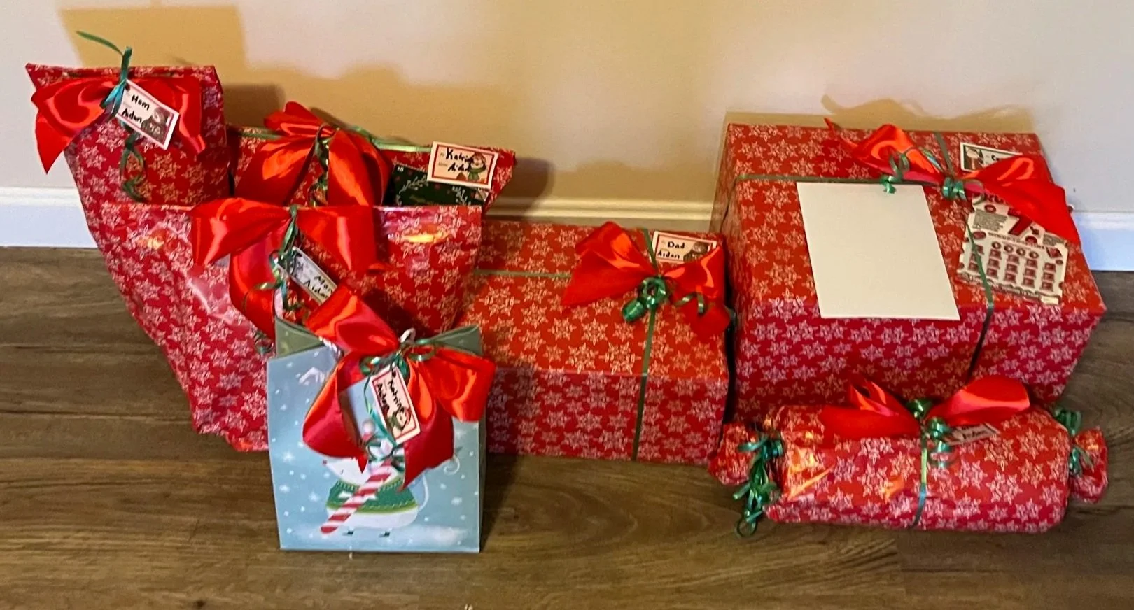 Multiple Christmas presents wrapped in red and white holiday paper, decorated with red bows and green ribbons, some with tags. One gift bag with a snowman and candy cane design.