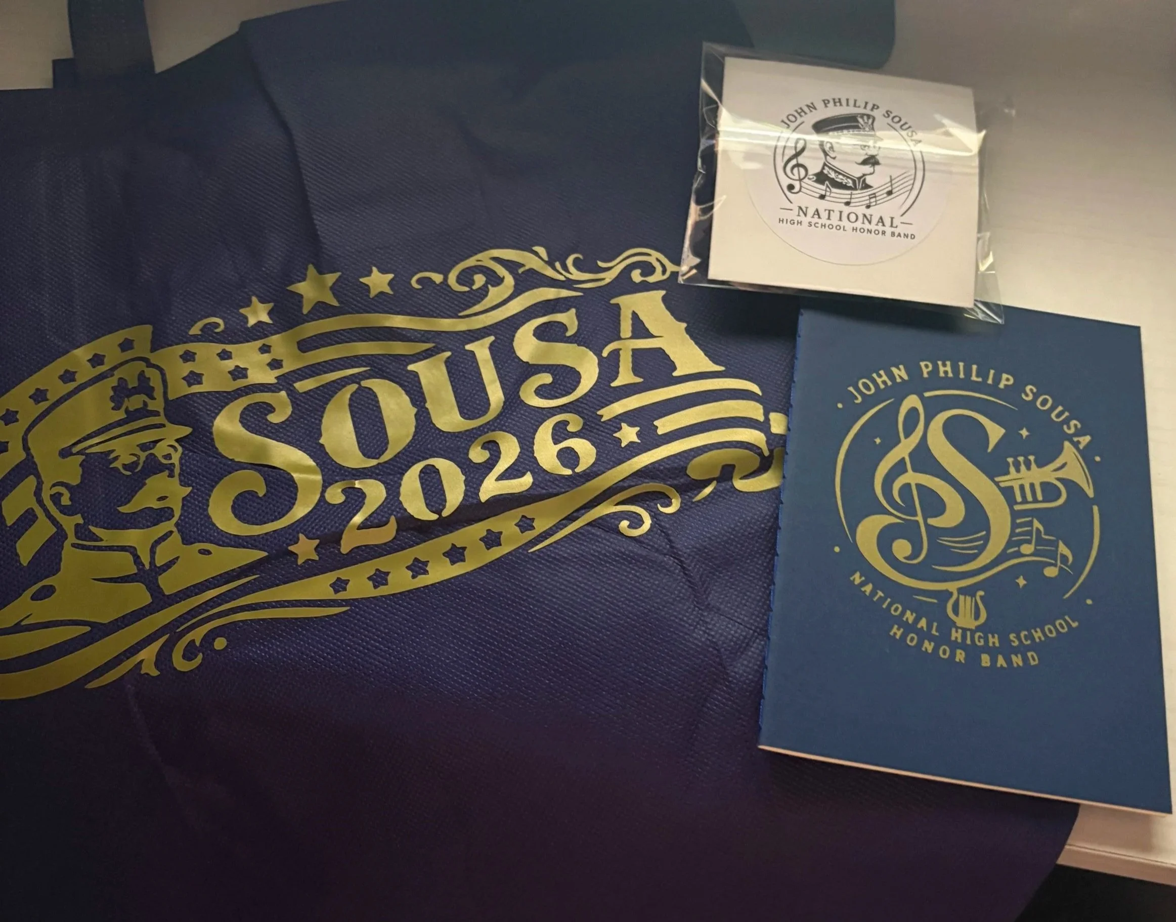 Close-up of a navy blue cloth with gold lettering and design, alongside a clear plastic package with a logo and text for the John Philip Sousa National High School Honor Band.