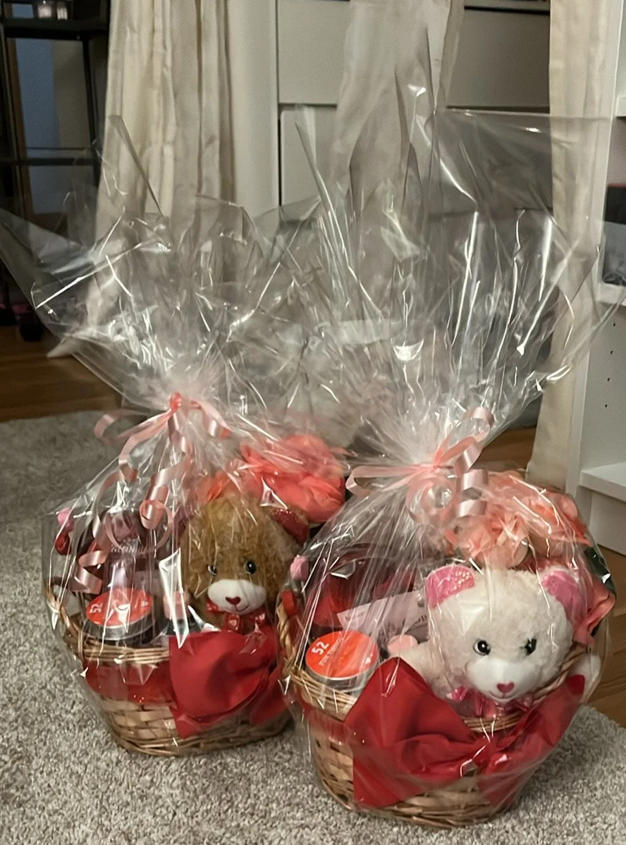 Two gift baskets wrapped in clear plastic, each decorated with pink ribbons. Inside are stuffed animal teddy bears, one brown and one white, surrounded by pink tissue paper.