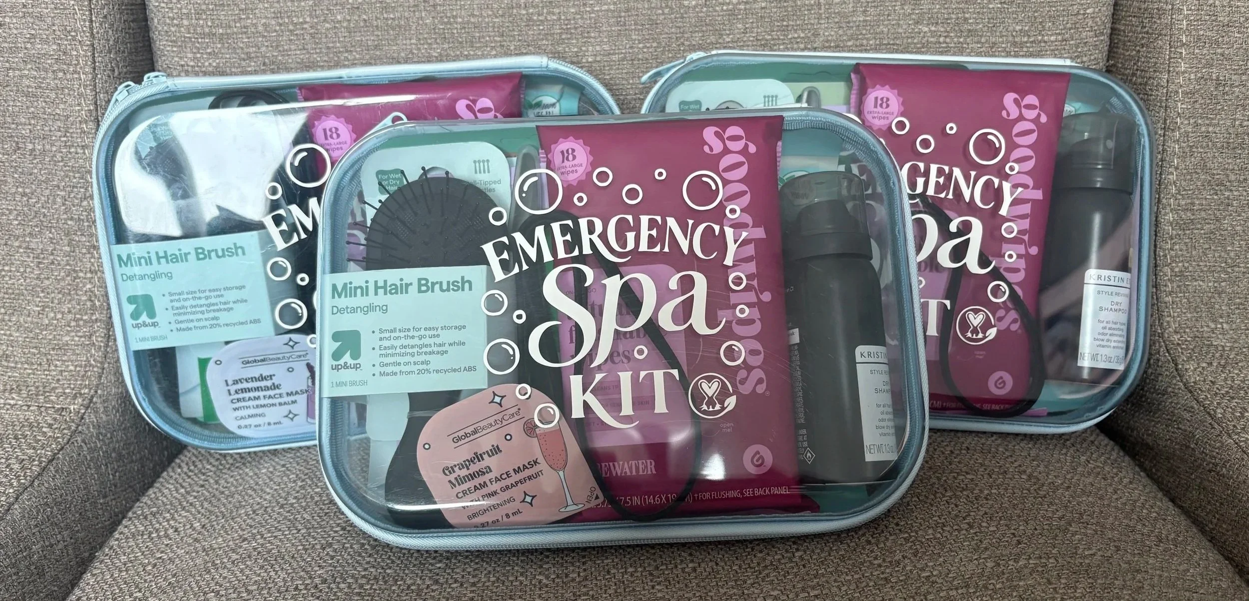 Three emergency spa kits with transparent zippered cases containing personal care items such as mini hair brushes, face masks, shampoo bottles, and wipes, placed on a brown fabric surface.