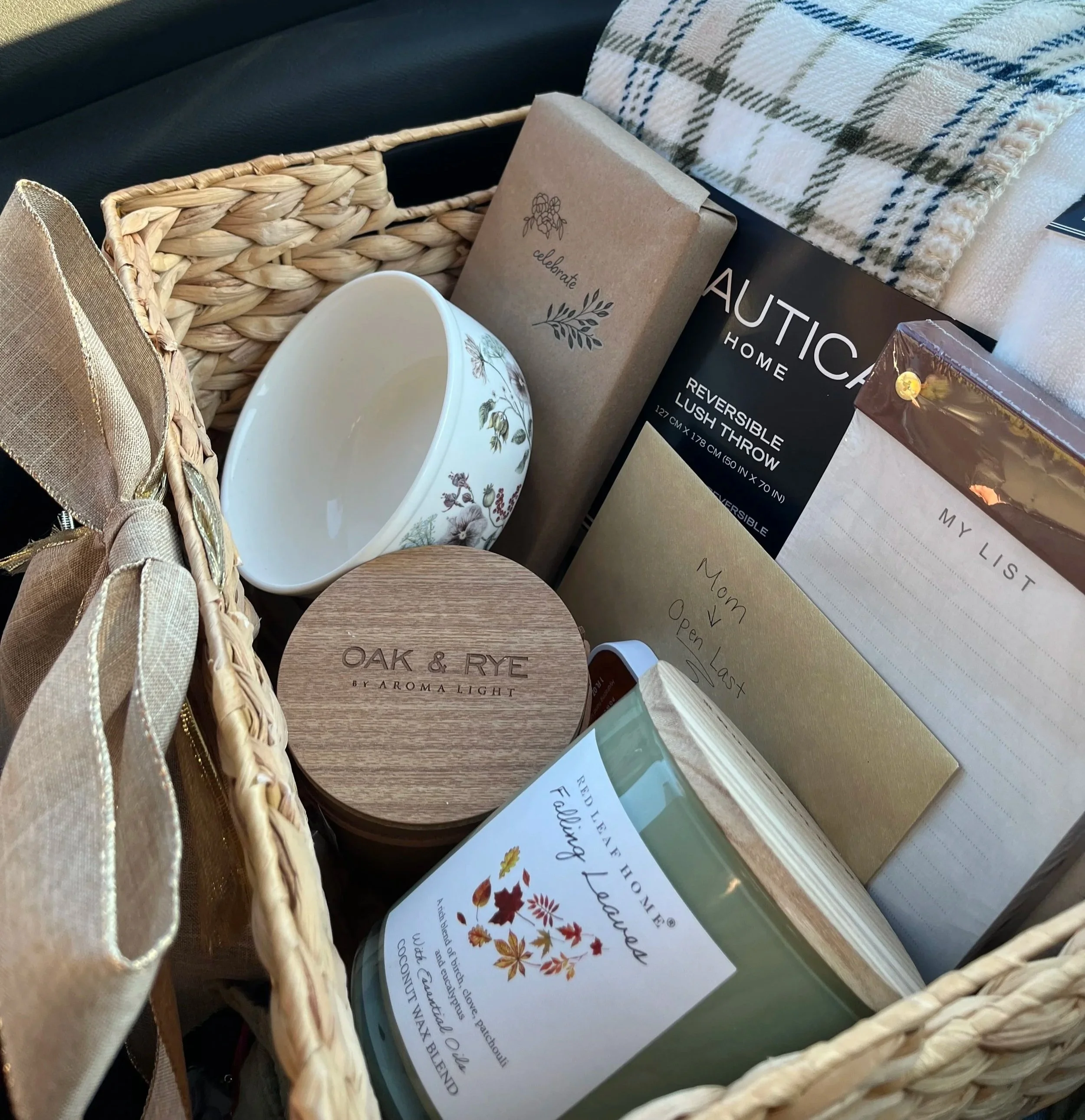 A wicker basket contains various household items including a ceramic mug with floral patterns, a brown paper bag with the word 'celebrate' and botanical illustrations, a box labeled 'OAK & RYE aroma light,' and a folded plaid cloth.
