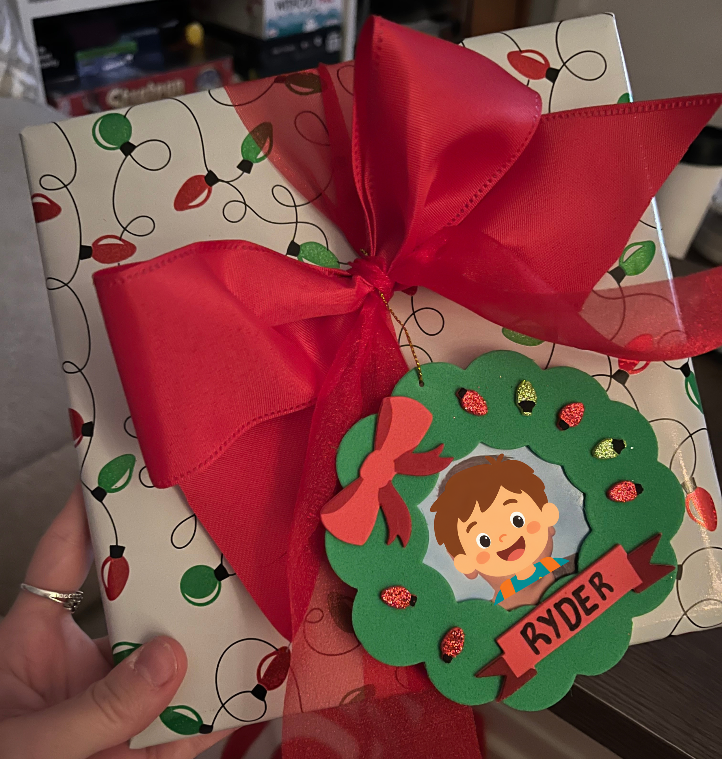 A wrapped Christmas gift decorated with holiday-themed wrapping paper, a large red bow, and a green paper tree ornament with glitter accents.