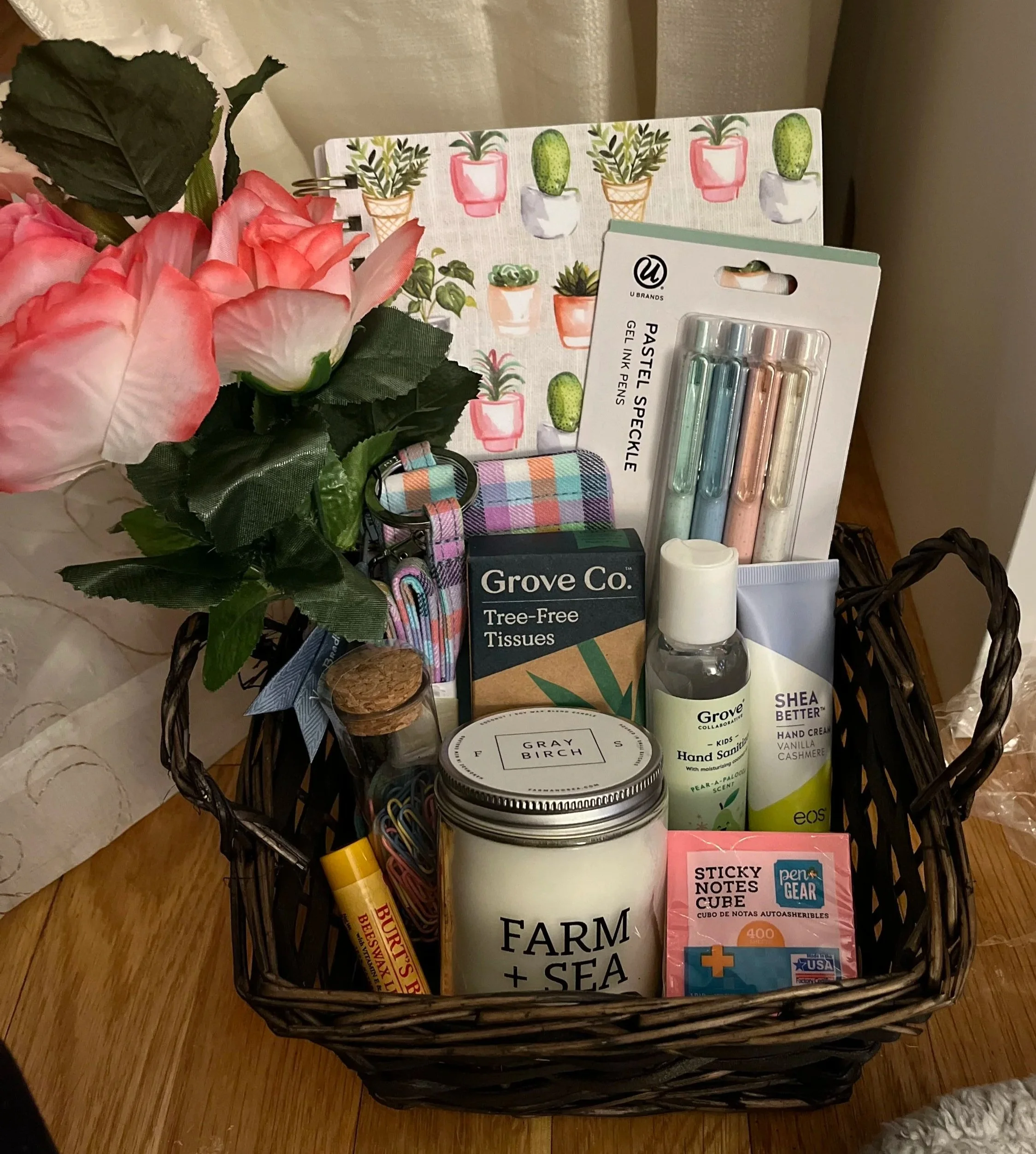 A black woven basket contains artificial pink and white flowers, and various office and personal items including a pack of gel ink pens, a box of tree-free tissues, a container of hand sanitizer, sticky notes, and a plaid fabric wristband. In the bac