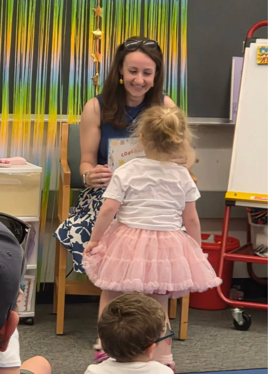 A woman sitting on a chair receiving a certificate from a young girl in a classroom decorated with colorful streamers and star-shaped decorations. The woman is smiling and wearing glasses on her head and yellow earrings. The girl is wearing a pink tutu skirt and a white shirt, with her back to the camera. There is a whiteboard and other classroom supplies visible in the background.