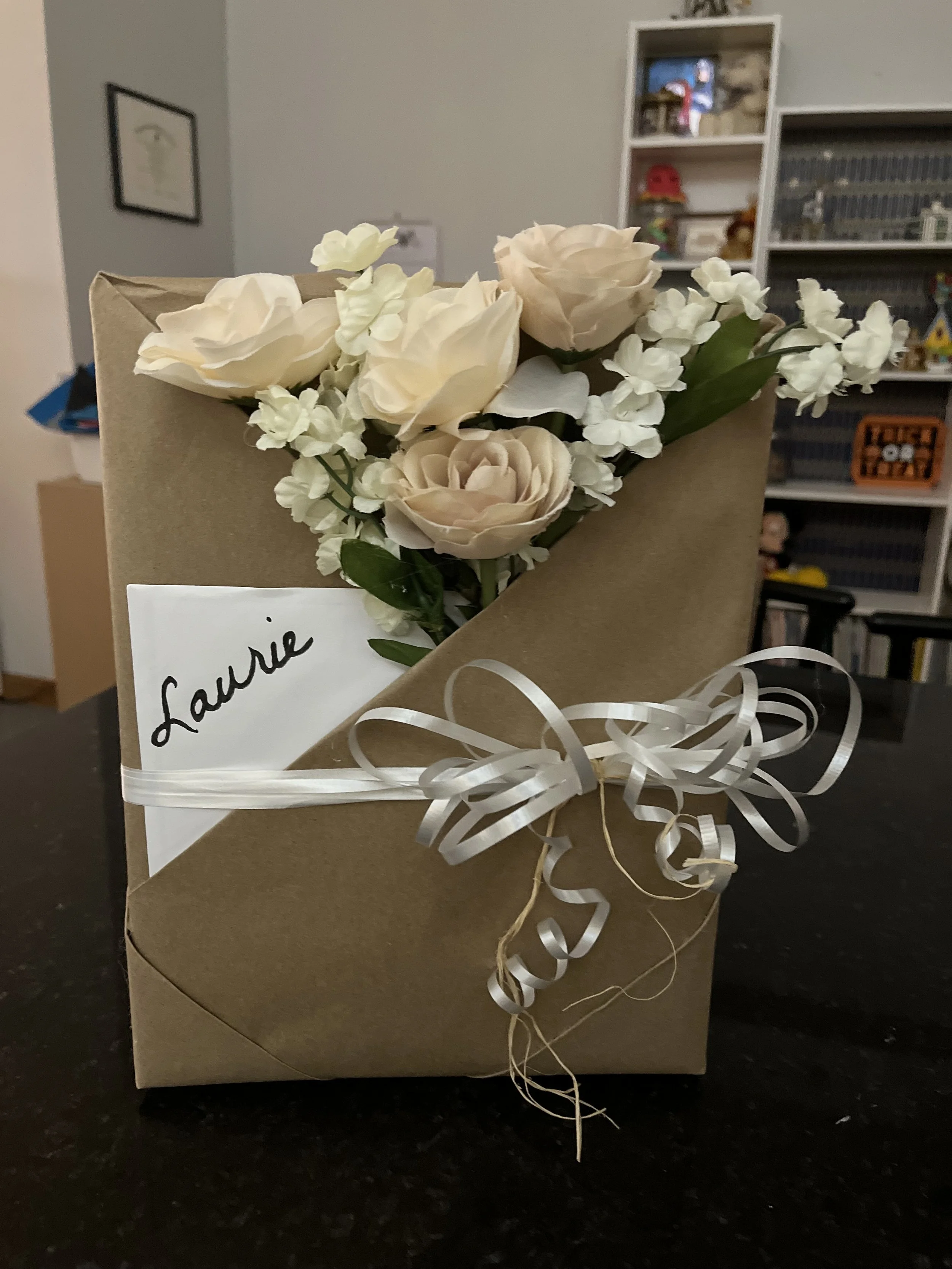 A cardboard gift box decorated with white flowers, including roses and small blossoms, along with green leaves, tied with white curled ribbon, and a handwritten note that says 'Laurae'.