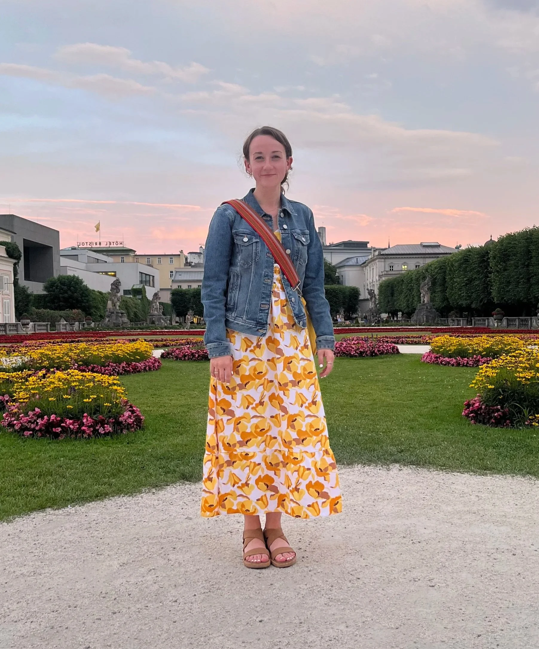 A woman stands on a gravel pathway in a park with colorful flower beds. She is wearing a long yellow dress with orange print, a blue denim jacket, and tan sandals. The sky is overcast with pinkish hues and the background features buildings and greenery.