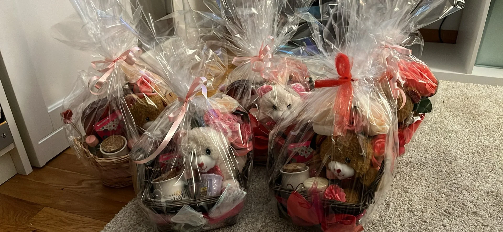 Three gift baskets wrapped in clear cellophane with pink and red ribbons, containing stuffed animals, mugs, and chocolates, on a wooden table and carpeted floor.
