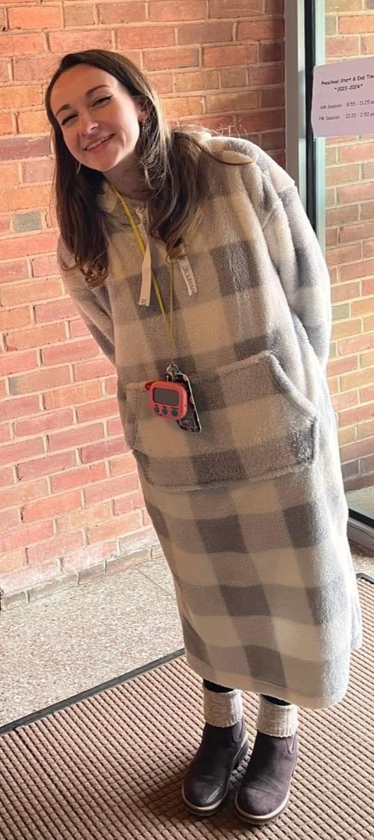 A young woman standing indoors near a glass door, smiling at the camera, wearing a long checkered fleece coat, black boots, and gray socks, with a brick wall in the background.