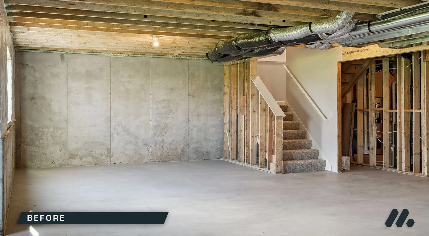 Empty basement with concrete floor, exposed ceiling beams, and ductwork, with a staircase and framing for walls.